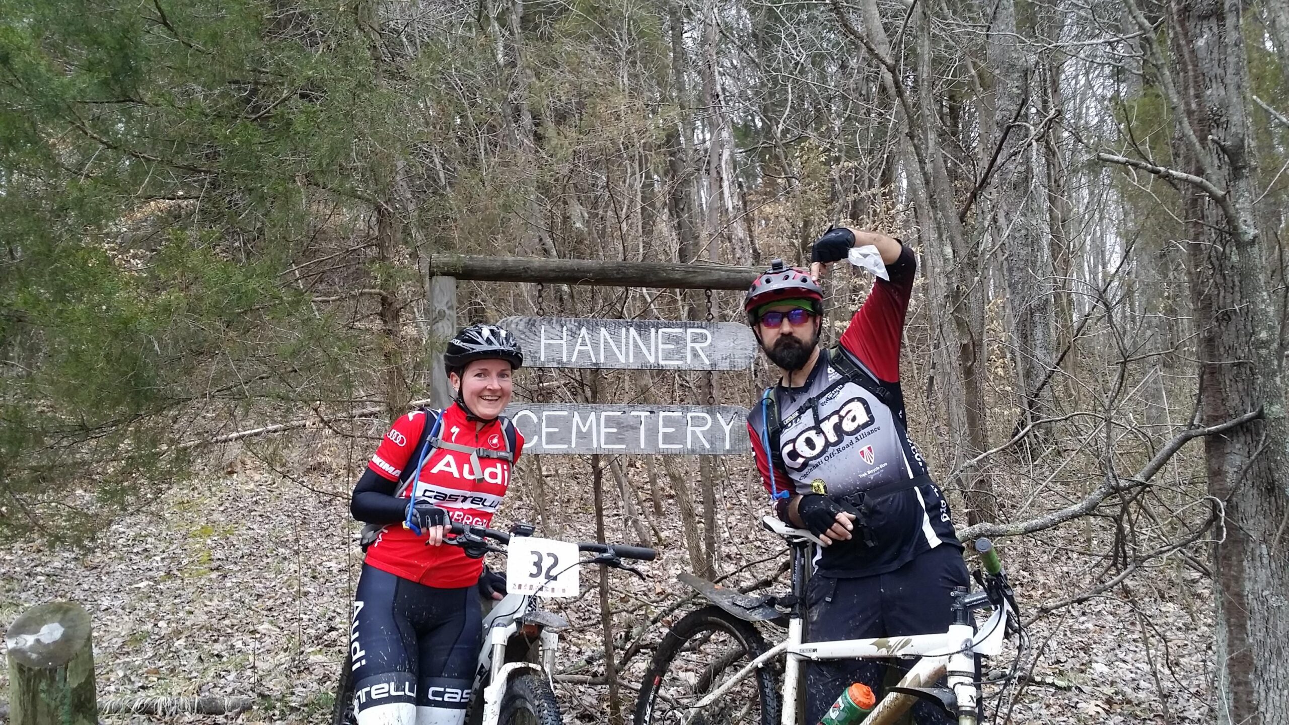 Two mountain bikers smile for the camera in front of a wooden sign that reads "Hanner Cemetery." They are dressed in cycling gear, with one person raising an arm playfully while the other stands beside a bike. The background features a wooded area with trees and fallen leaves. Hickory Ridge mountain bike trail.