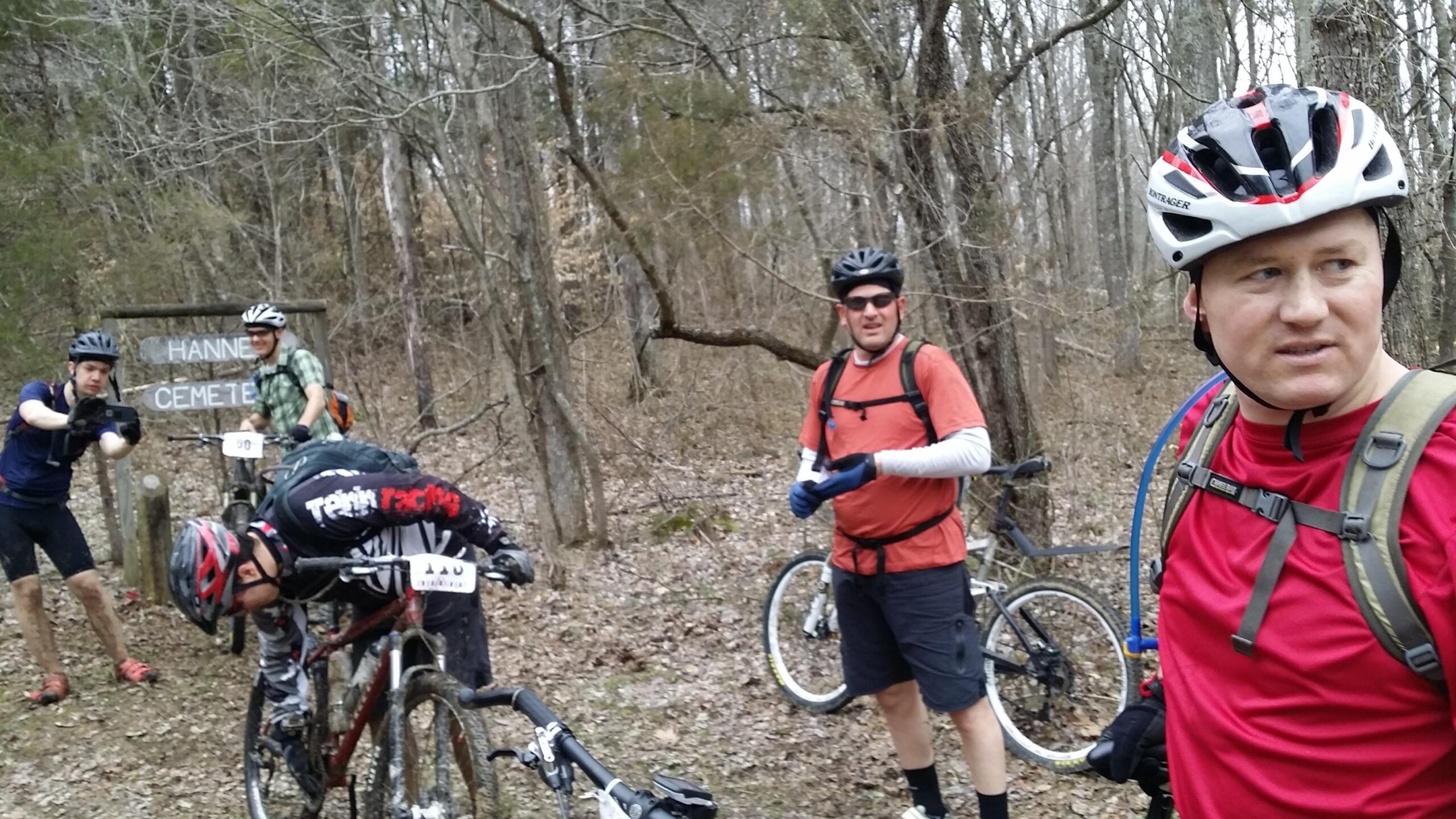 A group of mountain bikers, wearing helmets and cycling gear, gather near a woodland area with a sign that reads "Hanner Cemetery." Two cyclists are tending to their bikes while another takes a photo. The scene is surrounded by trees in a leaf-covered environment, suggesting a cool, early spring day. Hickory Ridge mountain bike trail.
