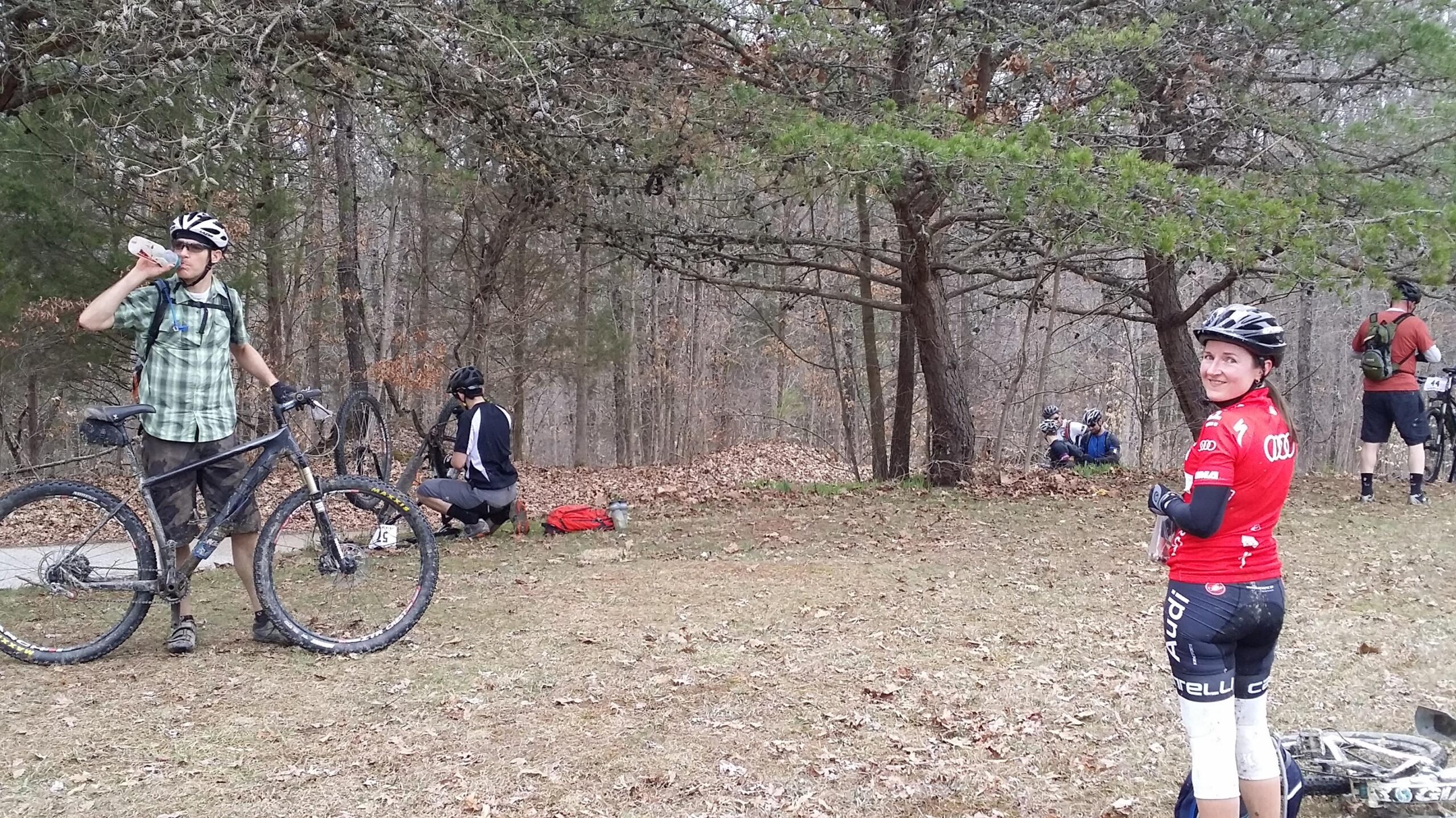 A group of mountain bikers resting in a wooded area. In the foreground, a man in a plaid shirt and helmet drinks from a water bottle while standing next to his bike. To his right, a woman in a red cycling jersey and helmet smiles at the camera. In the background, another cyclist is crouched near a bike, and additional riders can be seen seated on the ground. The scene is set among trees with fallen leaves on the ground. Hickory Ridge mountain bike trail.
