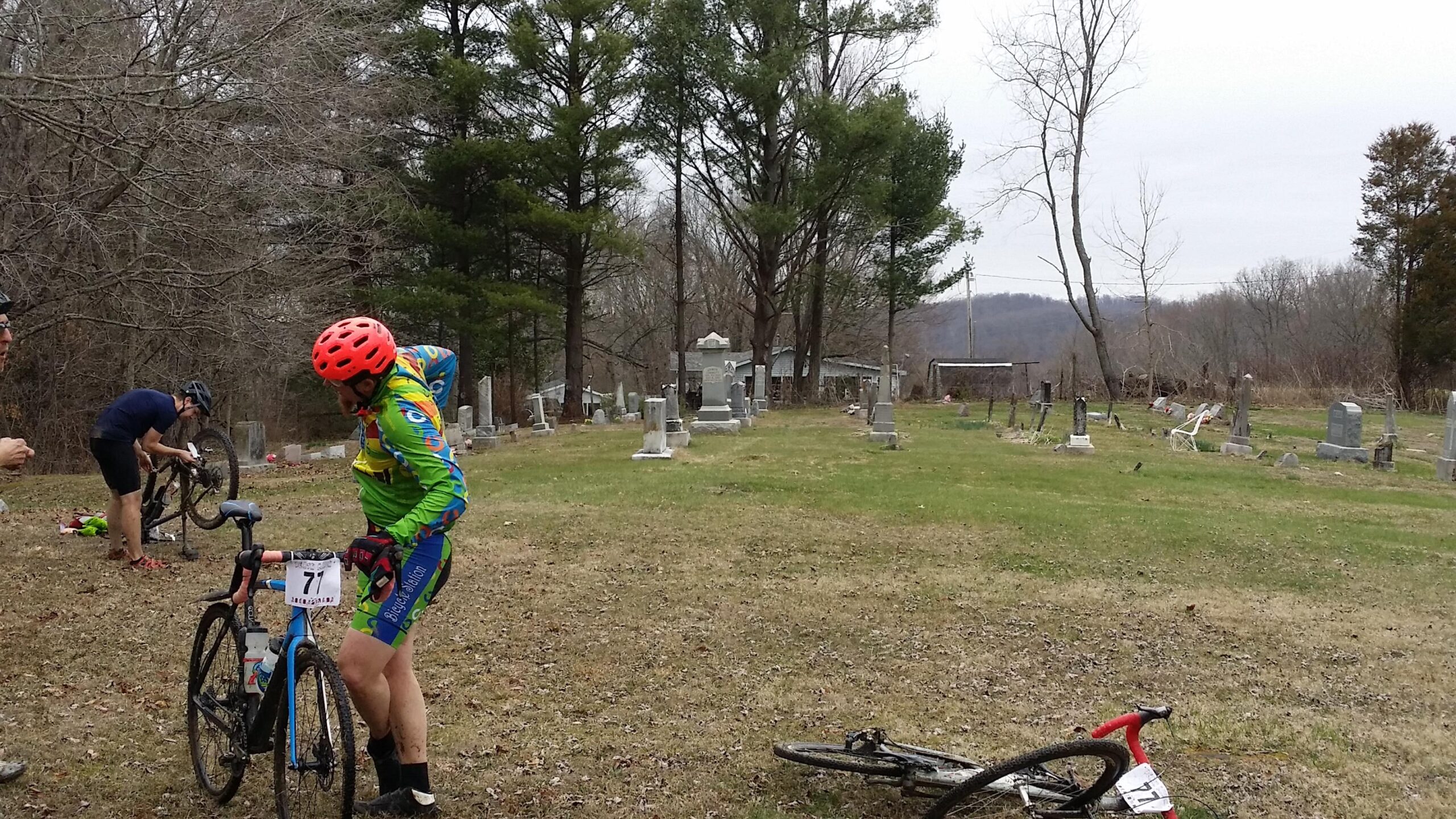 Two cyclists are seen in a grassy area near a cemetery. One cyclist, dressed in a bright green and blue outfit with a red helmet, is checking their bike. The other cyclist, in a dark shirt, is repairing a mountain bike. In the background, several tombstones and trees are visible, creating a serene yet unusual setting for cycling activities. Hickory Ridge mountain bike trail.