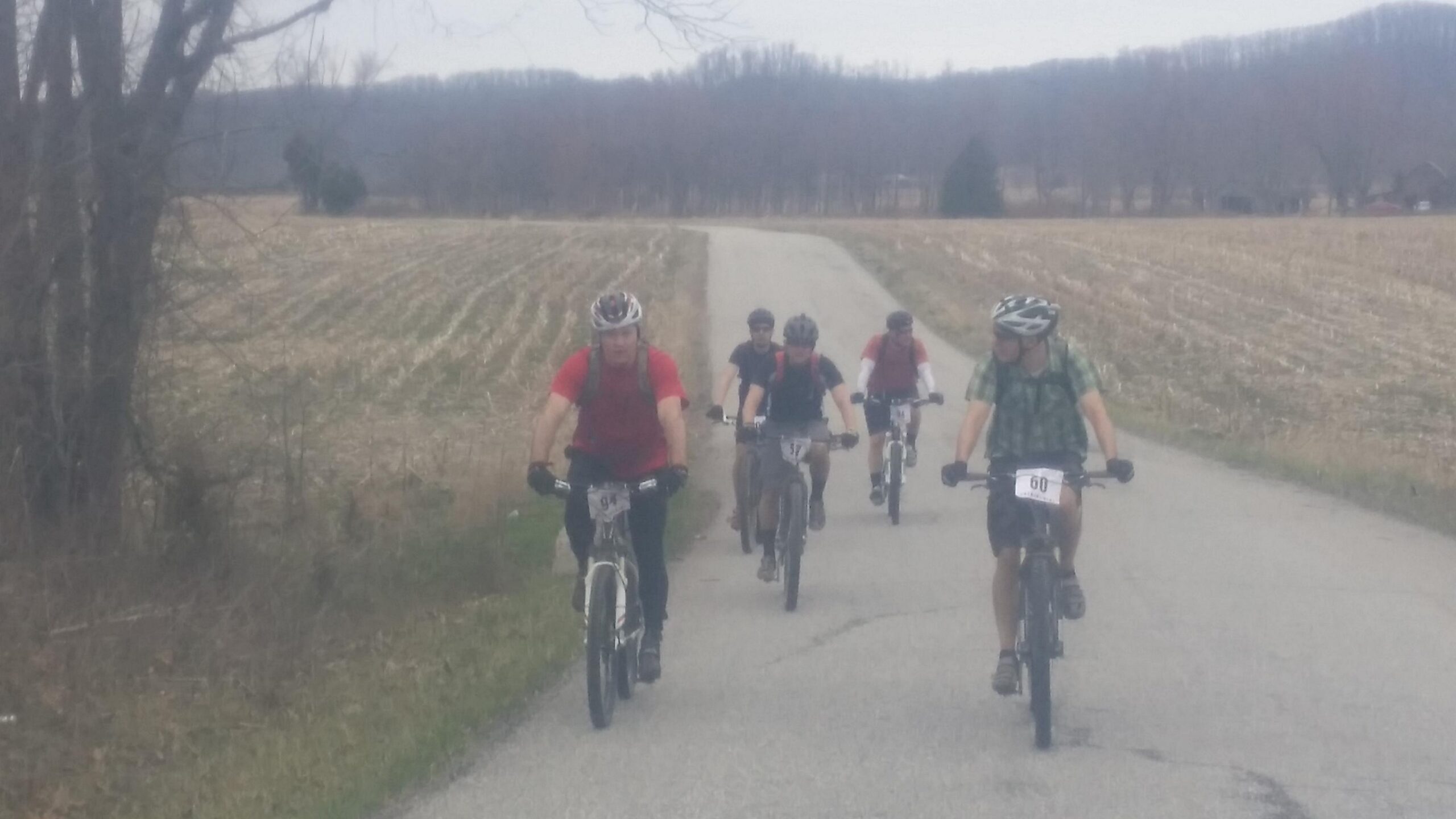 A group of five mountain bikers riding on a rural road, surrounded by fields and trees. The cyclists are wearing helmets and some have race numbers on their bikes. It's an overcast day, and the landscape features harvested fields in the background. Hickory Ridge mountain bike trail.