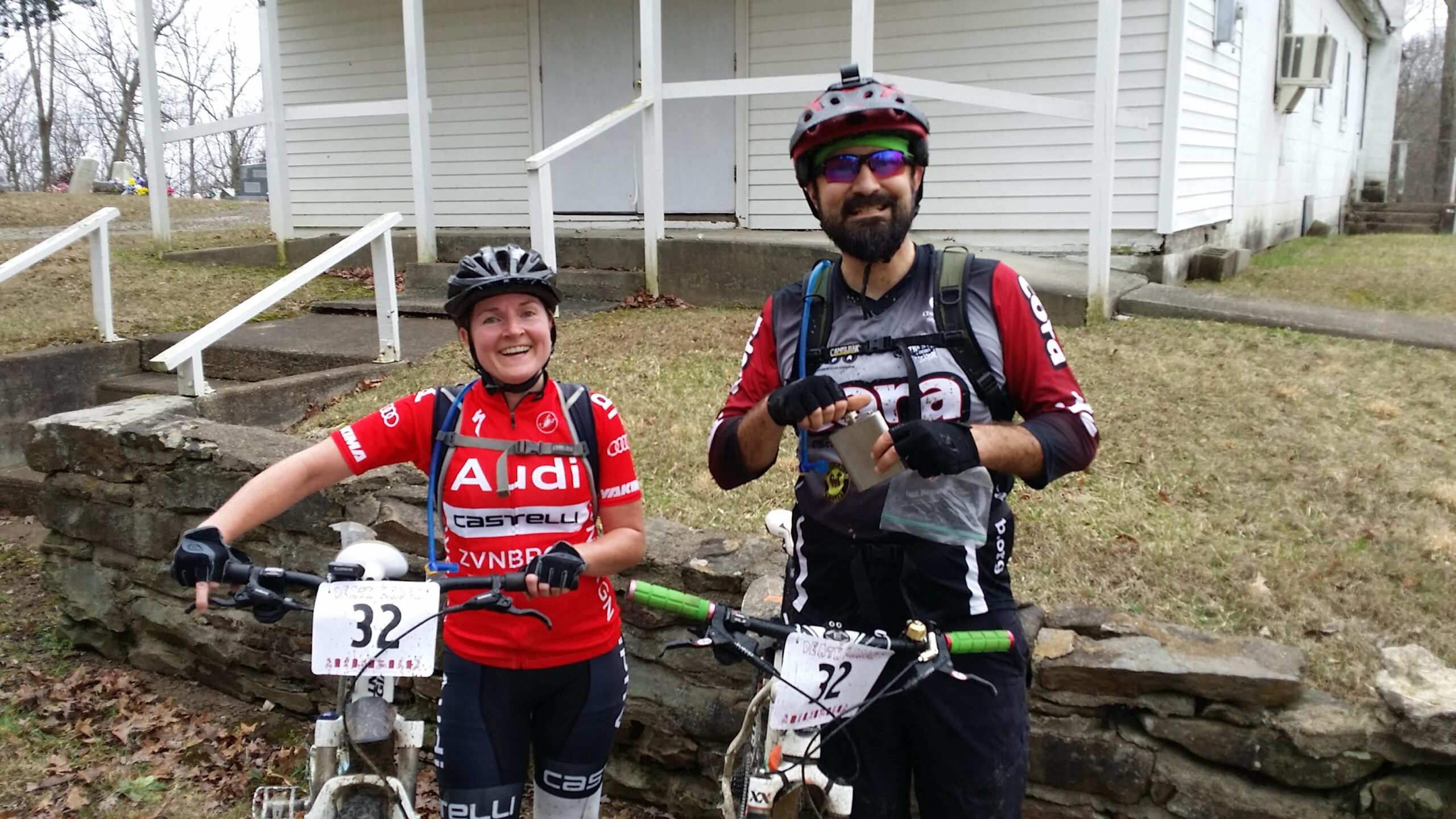 Two cyclists, a man and a woman, stand proudly next to their mountain bikes, smiling at the camera. The woman wears a red cycling jersey with logos and has a helmet on. The man, in a gray and red jersey, holds a small item in his hand. They are outdoors, with a white building and grassy area in the background. Hickory Ridge mountain bike trail.