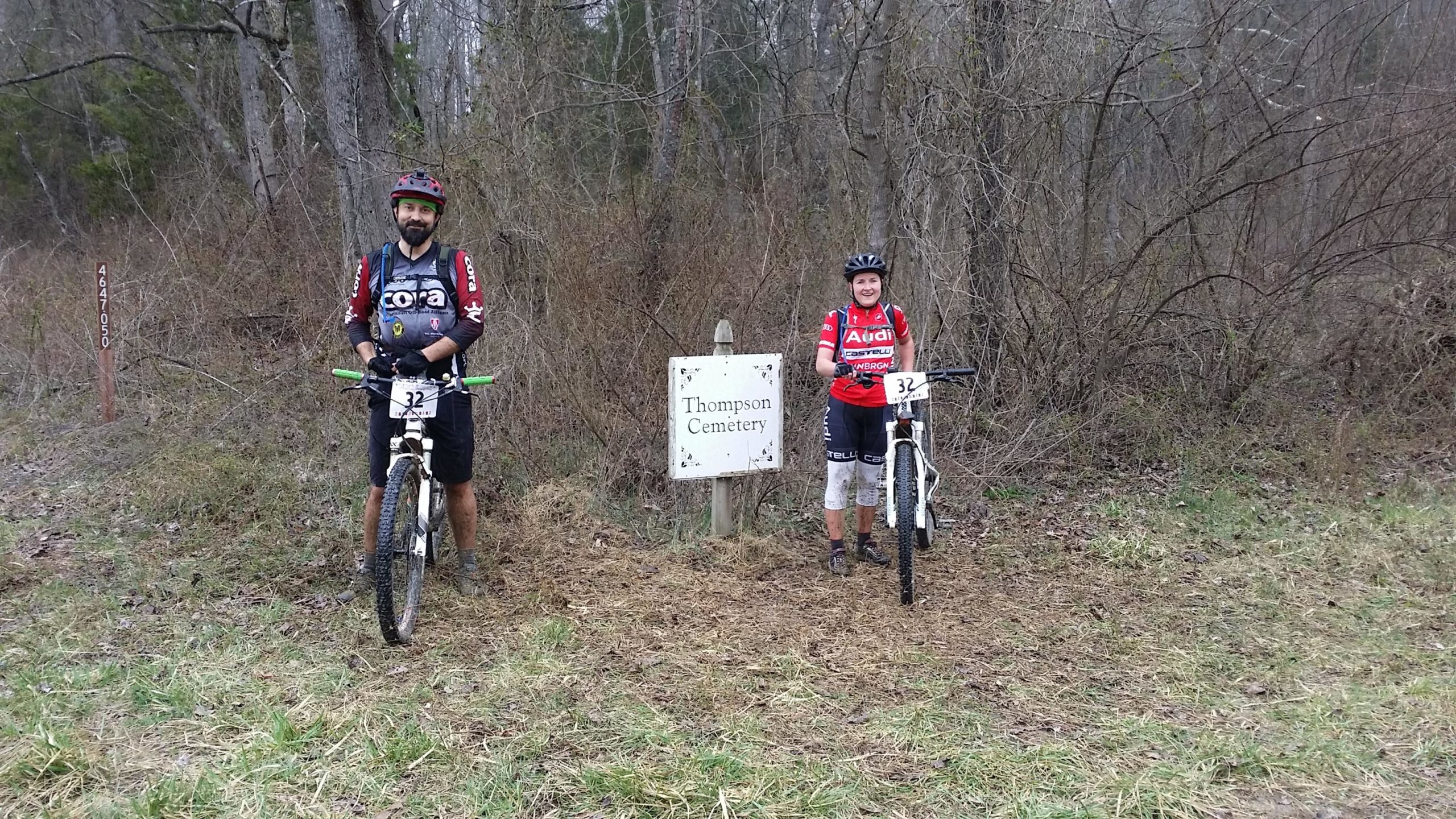 Two mountain bikers pose next to a sign for "Thompson Cemetery," surrounded by a wooded area with sparse vegetation. The male biker is on the left in a dark jersey and shorts, while the female biker is on the right in a red jersey and white pants. Both are wearing helmets and are positioned next to their bikes, which have race numbers attached. Hickory Ridge mountain bike trail.