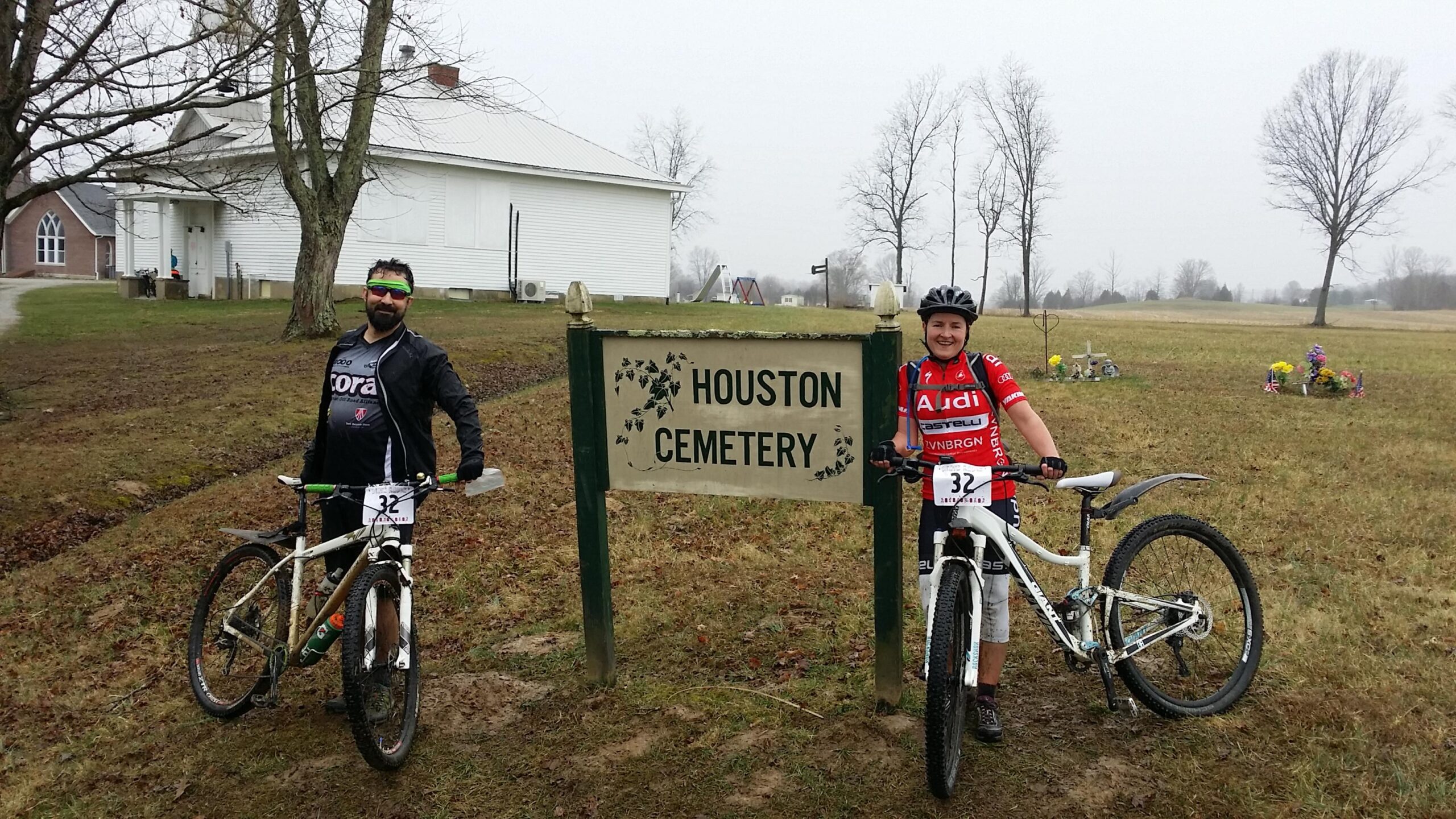 Two mountain bikers pose next to a sign that reads "Houston Cemetery." The scene is set in a grassy area with a few trees and a cloudy sky. One rider is wearing a black jersey and sunglasses, while the other is in a red jersey with logos. Both are smiling and leaning on their bikes, which are positioned beside the sign. Hickory Ridge mountain bike trail.