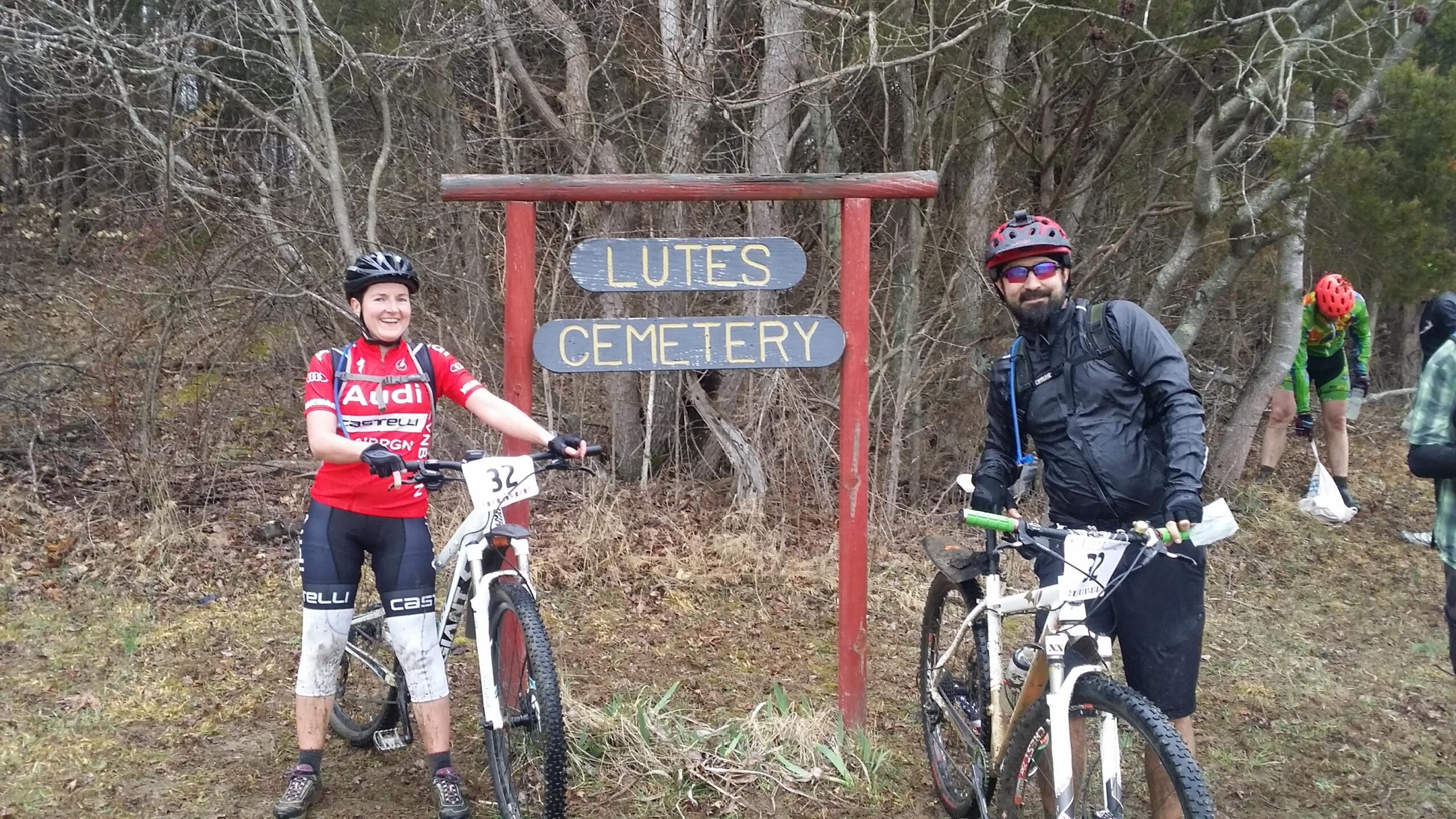 Two mountain bikers are posing in front of a sign that reads "Lutes Cemetery." The woman on the left is wearing a red cycling jersey and has a big smile, while the man on the right is wearing a black jacket and sunglasses. Both are beside their bikes, and the background features a wooded area. Some other cyclists are partially visible behind them. Hickory Ridge mountain bike trail.