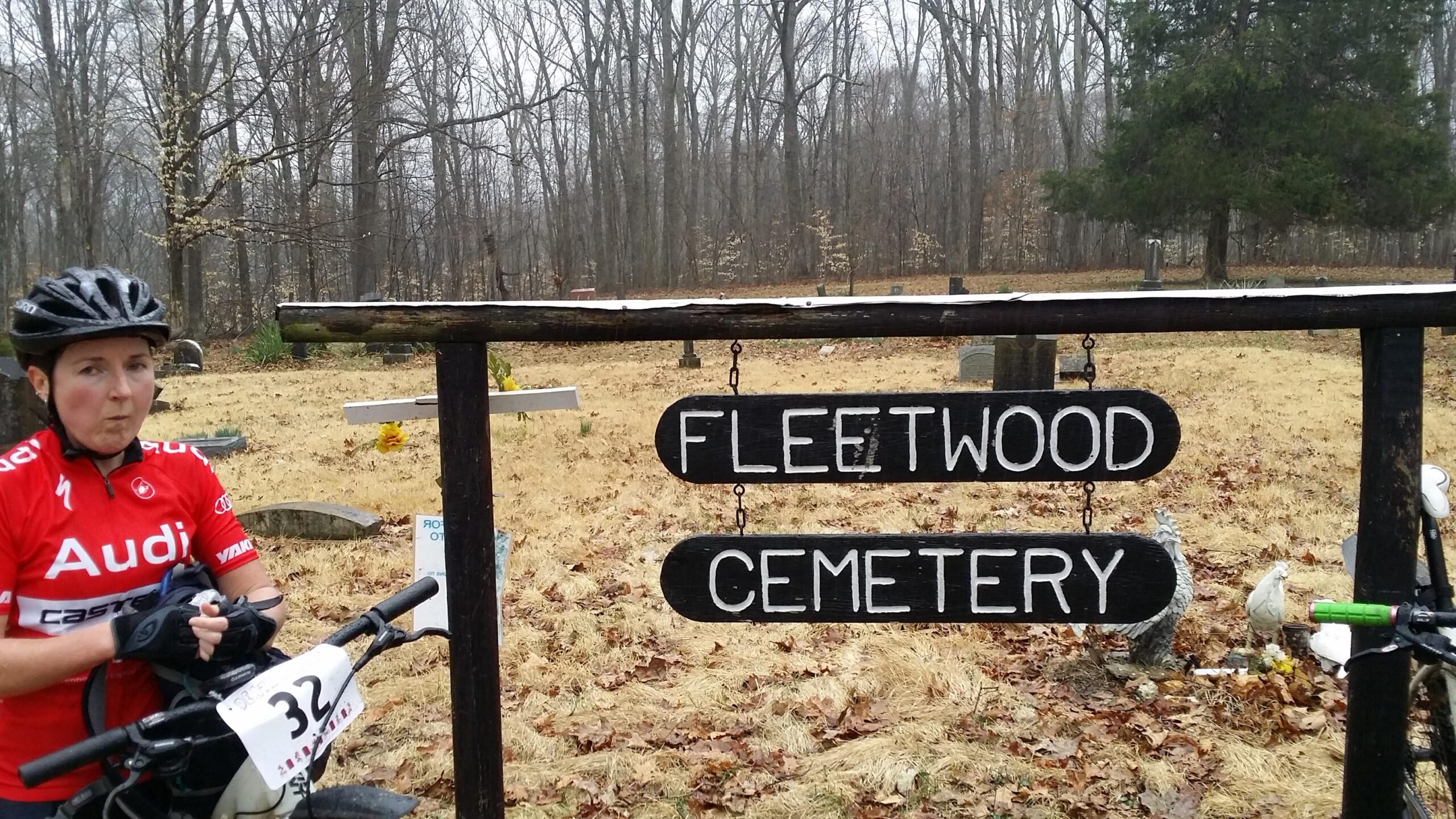 A cyclist in a red jersey stands beside a wooden sign that reads "Fleetwood Cemetery." The background features a cemetery with gravestones surrounded by bare trees and dry grass, suggesting a chilly, overcast day. The cyclist appears to be taking a break, holding their bike, and looking directly at the camera. Hickory Ridge mountain bike trail.