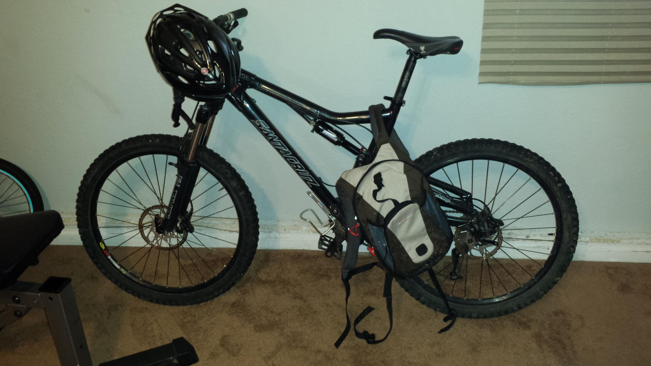 Santa Cruz Heckler: A black mountain bike with thick tires and a helmet resting on the handlebars, paired with a brown and gray backpack hanging from the frame, set against a light-colored wall and carpet.
