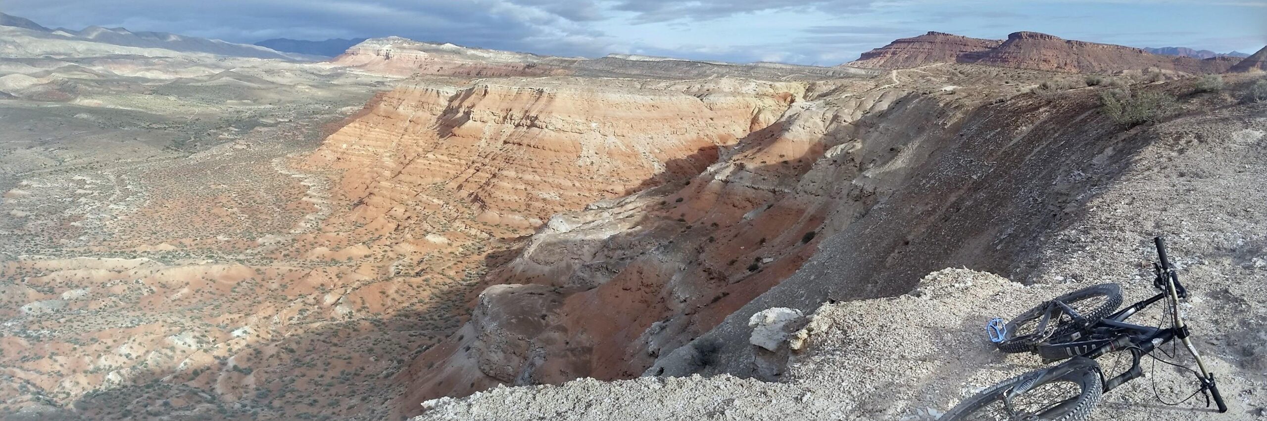 Intense Tracer 275: A panoramic view of a rugged desert landscape featuring steep cliffs and rolling hills in shades of orange and beige. In the foreground, a mountain bike lies on the edge of a rocky outcrop, overlooking the vast terrain. The sky above is partly cloudy, adding texture to the scene.