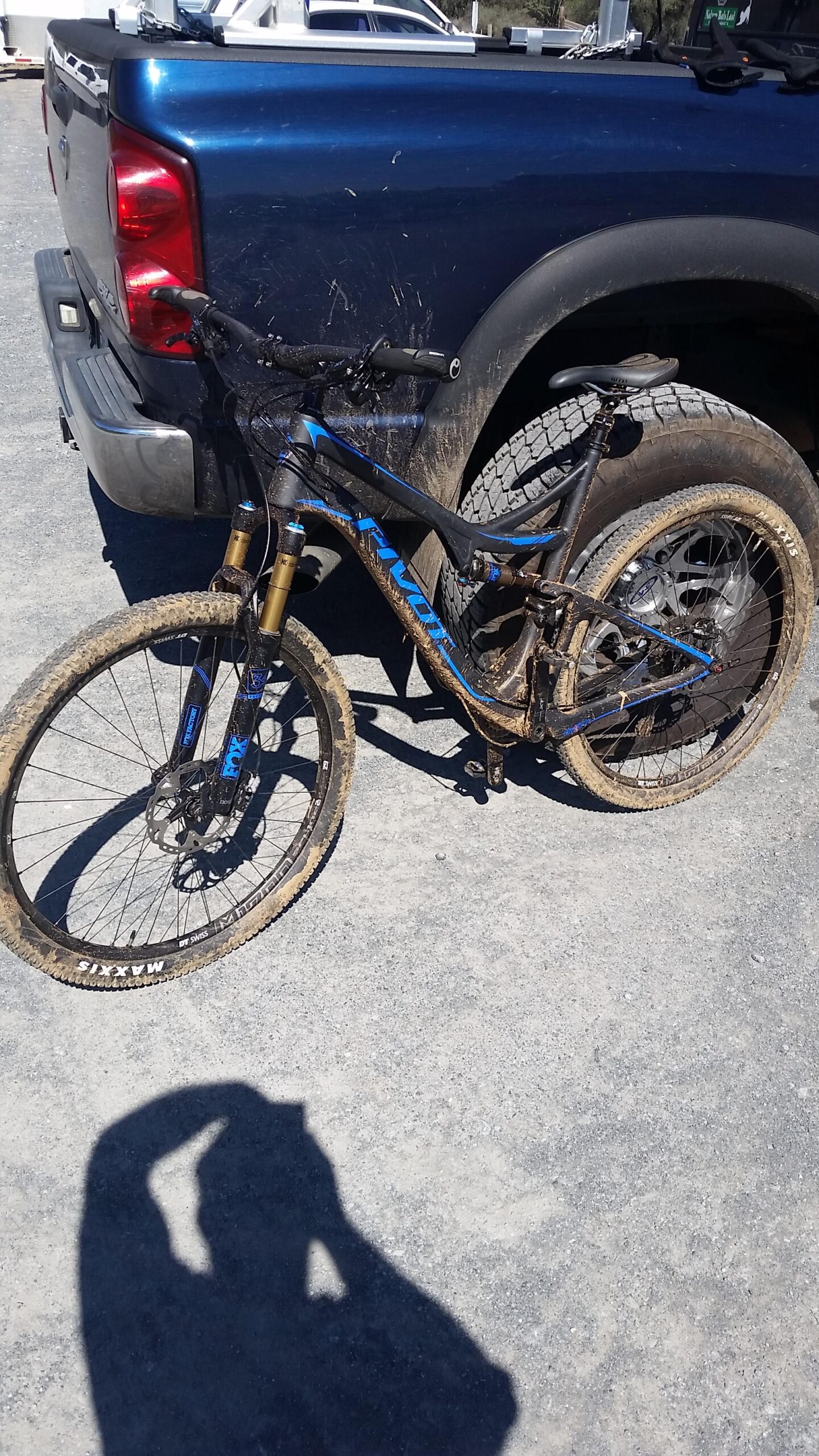 Pivot Mach 429 Carbon: A muddy mountain bike leaning against a blue pickup truck, with dirt splashes visible on both the bike and the truck. The ground is gravelly, and the sun casts a shadow of the photographer taking the picture.