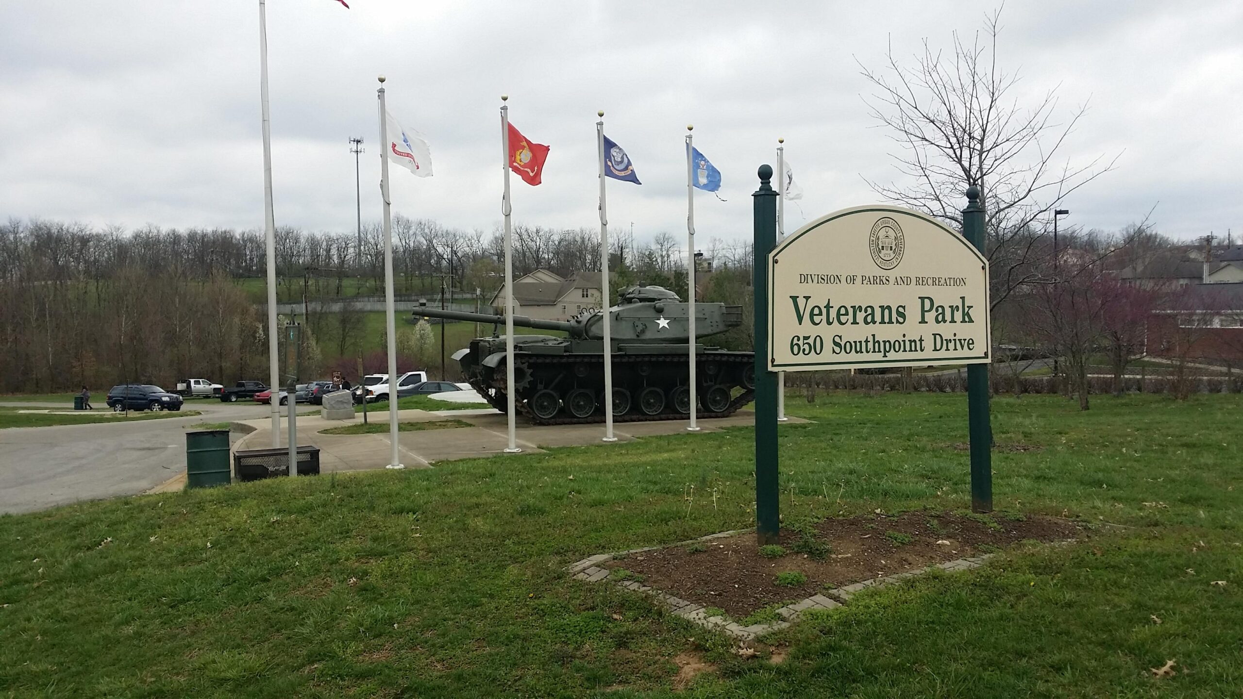 A grassy area featuring a sign for Veterans Park, which includes the address "650 Southpoint Drive." Nearby, several flags representing different military branches are displayed, and a military tank is positioned prominently in the background. The scene is set against a cloudy sky with trees and vehicles visible. Veterans Park mountain bike trail.