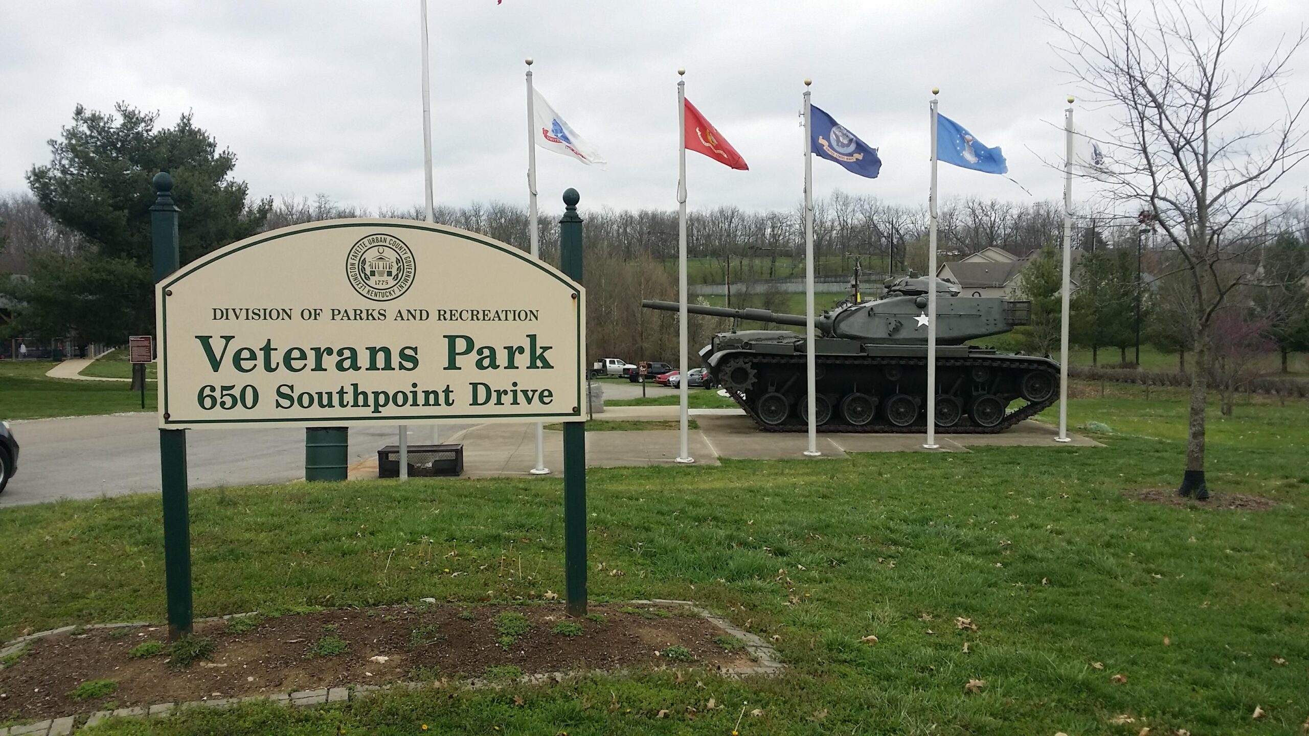 Sign at the entrance of Veterans Park displaying the name and address, with a military tank and several flags in the background, set against a cloudy sky. Veterans Park mountain bike trail.