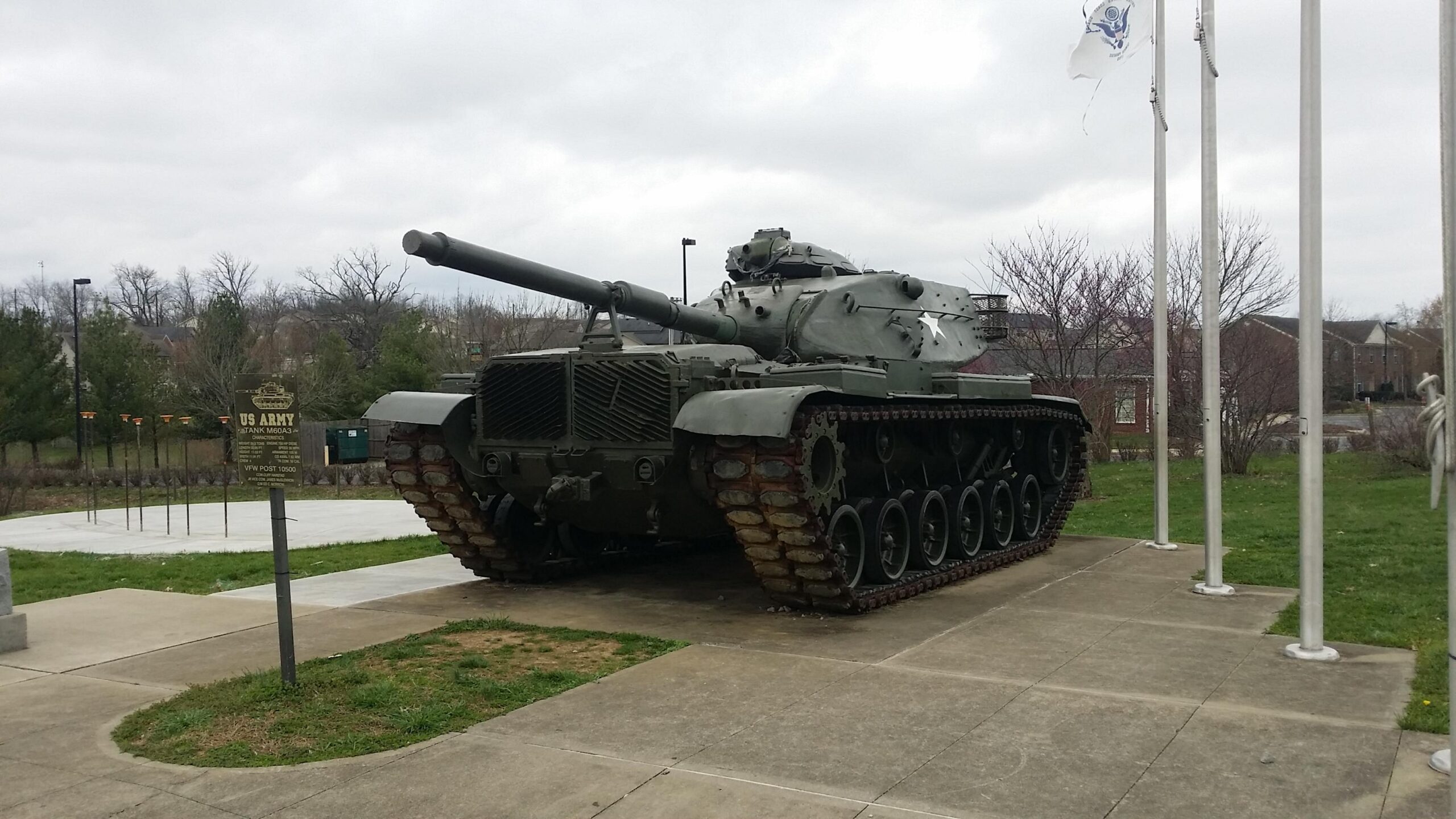 A military tank displayed outdoors on a concrete pad, with a sign indicating it is a US Army M60A3 tank. The tank is in a green camouflage color and has a long barrel. In the background, there are trees and flagpoles with flags, under a cloudy sky. Veterans Park mountain bike trail.