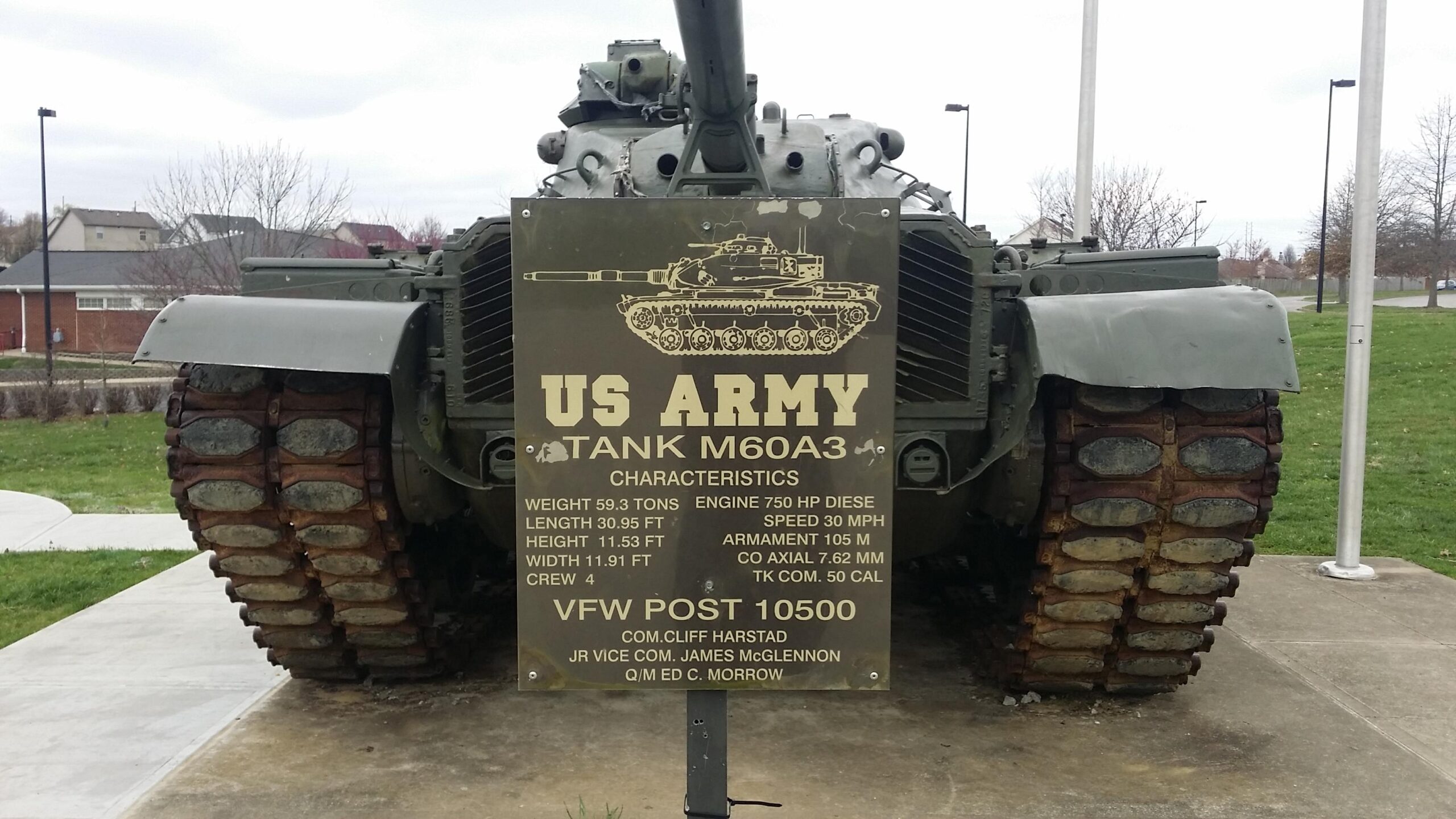 An M60A3 tank on display with a plaque detailing its specifications, including weight, length, height, engine horsepower, speed, and armament. The tank is set in a green park area with residential buildings in the background. Veterans Park mountain bike trail.