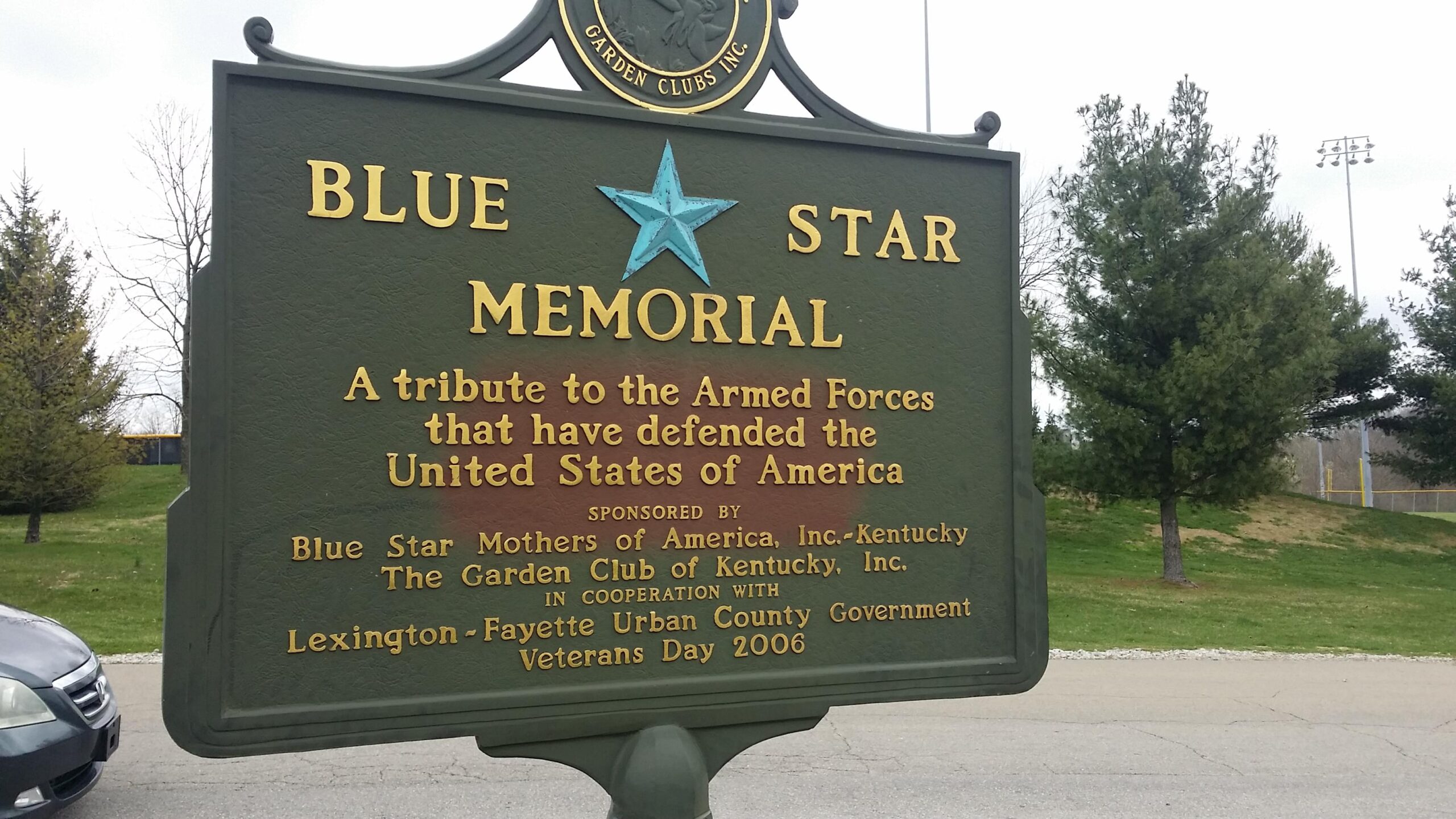 A large green sign reading "Blue Star Memorial" with a blue star emblem at the center. The sign honors the Armed Forces of the United States and mentions sponsorship by the Blue Star Mothers of America, the Garden Club of Kentucky, and the Lexington-Fayette Urban County Government, marked for Veterans Day 2006. Surrounding the sign are trees and a roadway. Veterans Park mountain bike trail.