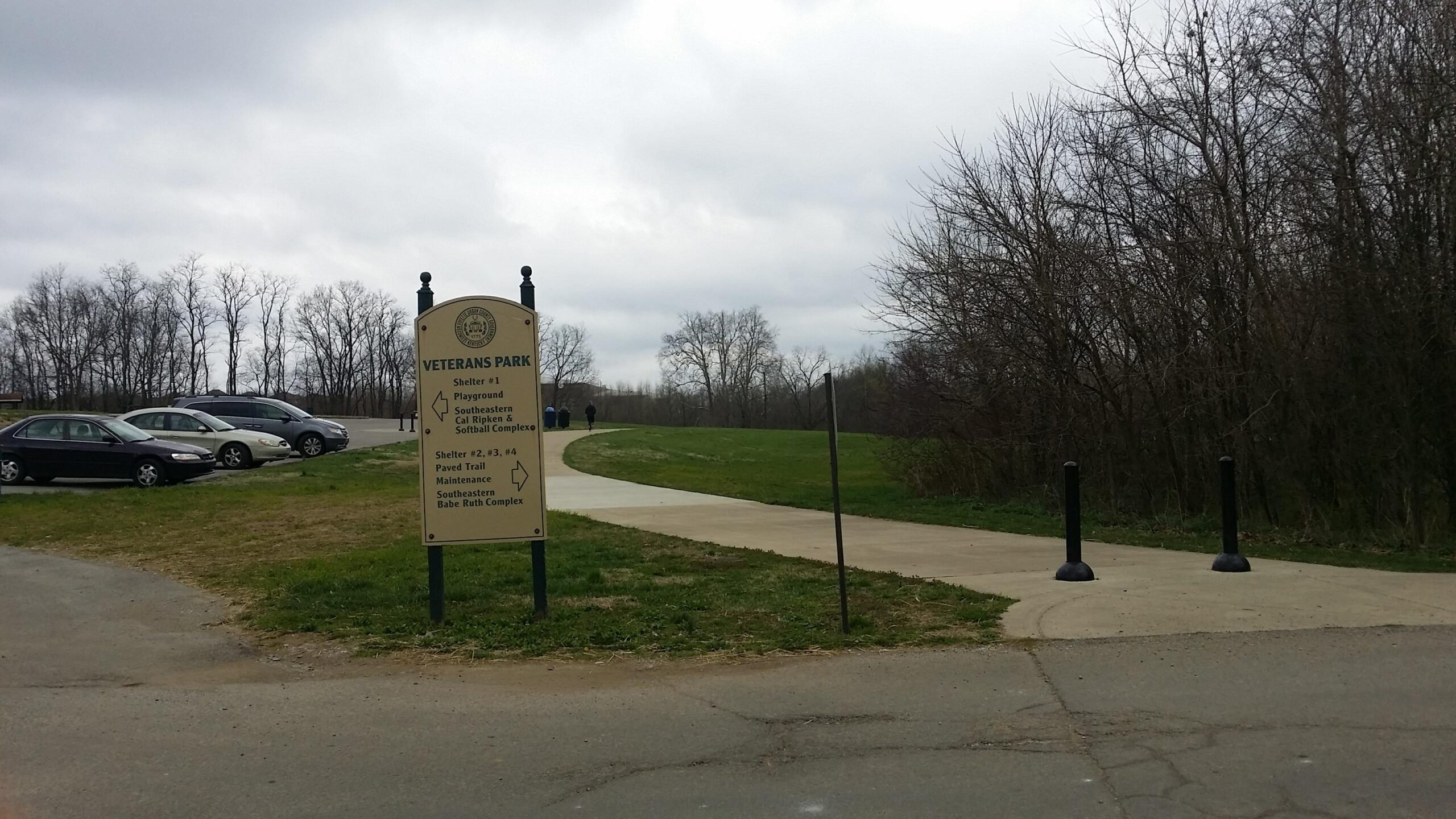 Sign for Veterans Park displaying directions to various amenities, including playgrounds, shelters, and a paved trail, with a parking area in the foreground and a cloudy sky overhead. Veterans Park mountain bike trail.
