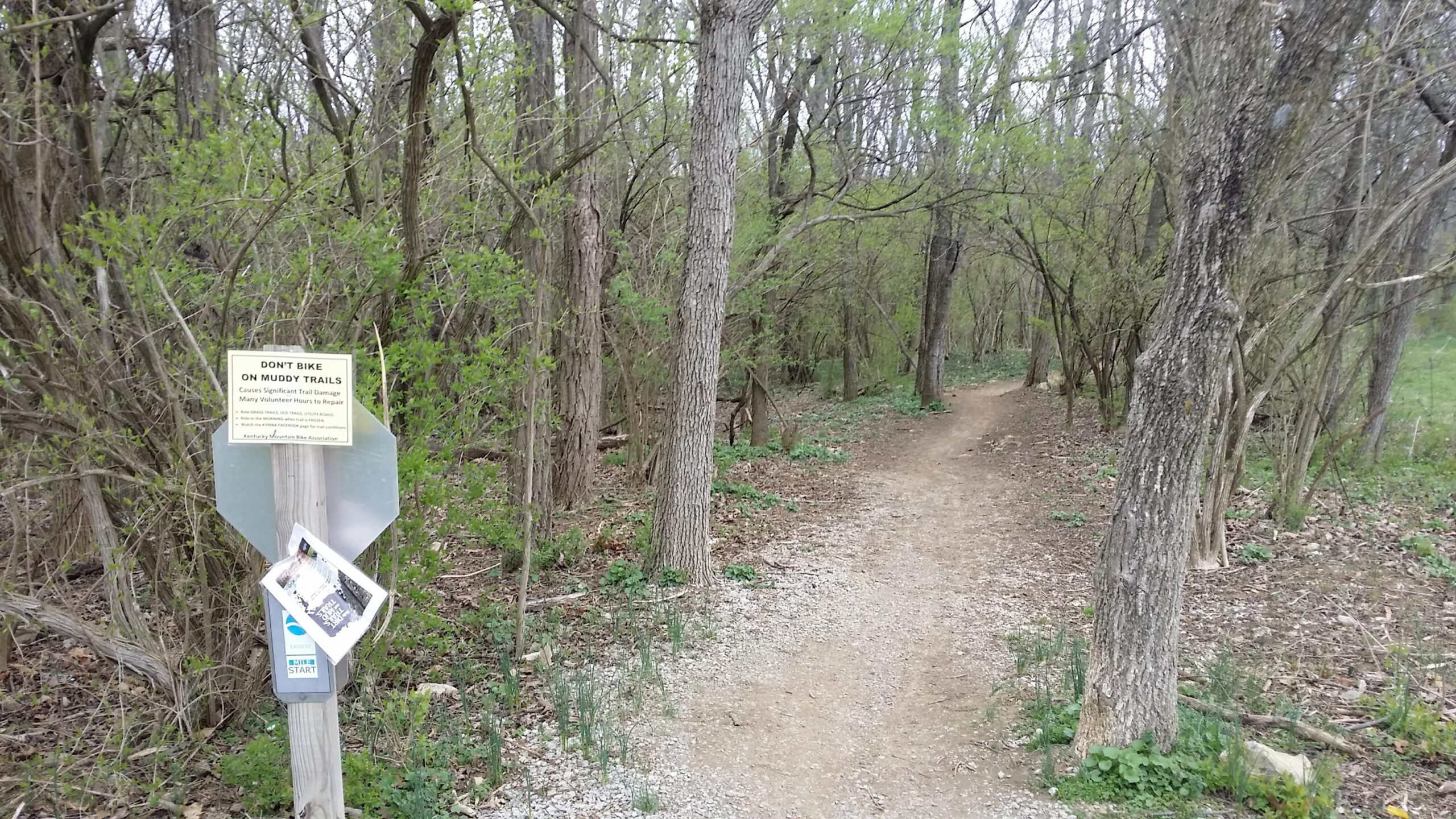 A wooded trail with a gravel path surrounded by trees and bushes. A sign at the trailhead reads "Don't bike on muddy trails" and provides information about trail care. The scene is green and lush, indicating early spring or late winter. Veterans Park mountain bike trail.