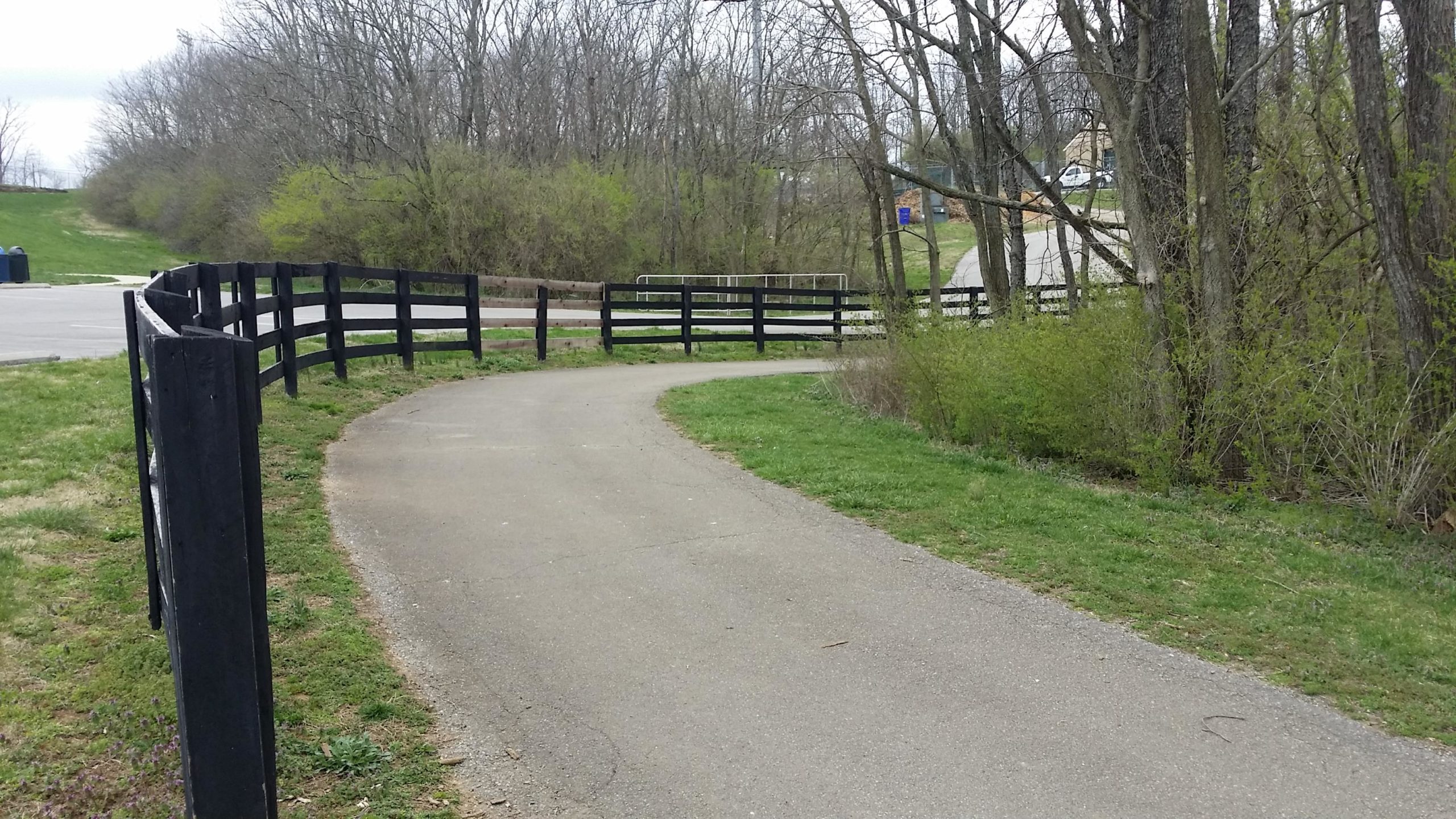 A winding paved path bordered by black wooden fencing, surrounded by trees and greenery, leading through a natural landscape. In the background, a glimpse of a parking area and a road can be seen. The scene is set on a cloudy day, adding a tranquil atmosphere. Veterans Park mountain bike trail.