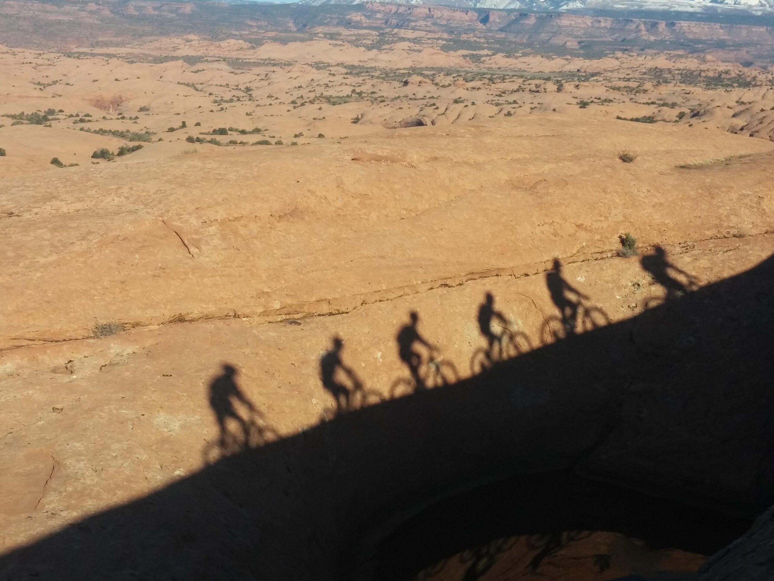 Silhouettes of four mountain bikers cast on a rocky landscape, showcasing a desert environment with distant hills and vegetation under a clear sky. Slickrock mountain bike trail.