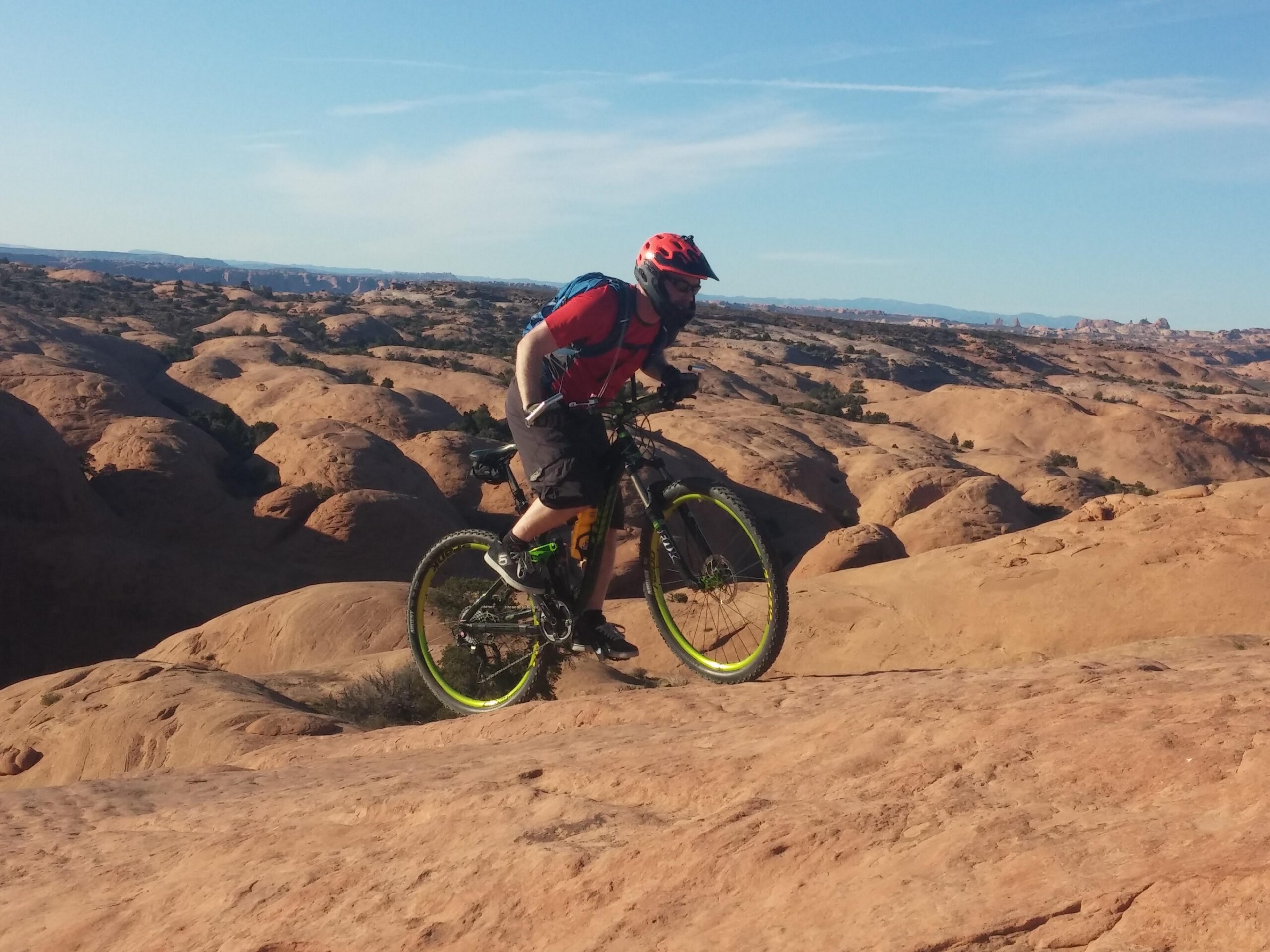 A mountain biker navigating rocky terrain on a sunny day, with expansive desert landscapes and blue sky in the background. The cyclist is wearing a helmet and sports gear, showcasing an adventurous outdoor activity. Slickrock mountain bike trail.