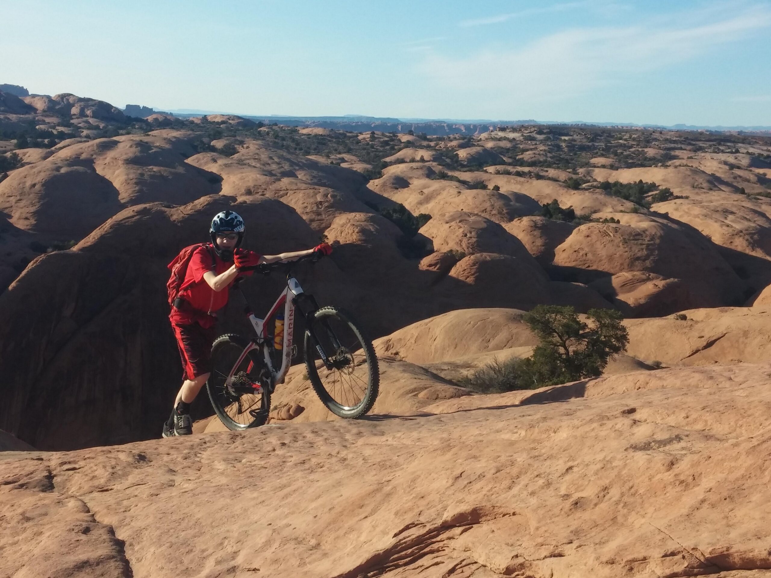 A person in red biking gear stands on a rocky terrain with a mountain bike, posing for the camera. The background features rugged, rounded rock formations under a clear blue sky. The scene captures the adventurous spirit of mountain biking in a natural landscape. Slickrock mountain bike trail.