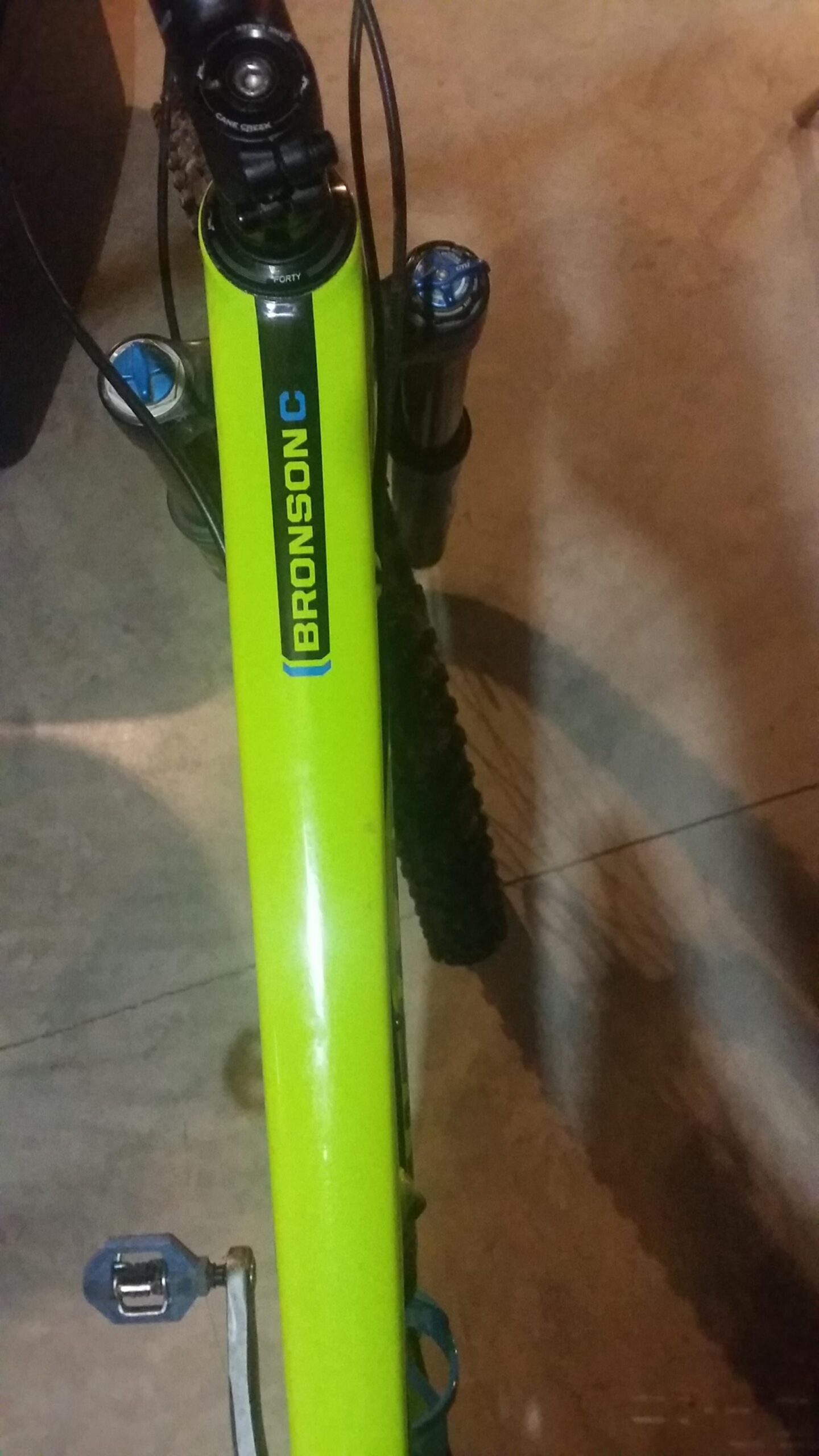 Santa Cruz Bronson: A close-up view of a bright green mountain bike frame with the name "BRONSON C" visible along the top tube. The image shows part of the bike's front suspension and features a visible pedal on the left side. The background is slightly blurred, suggesting the bike is indoors on a concrete surface.