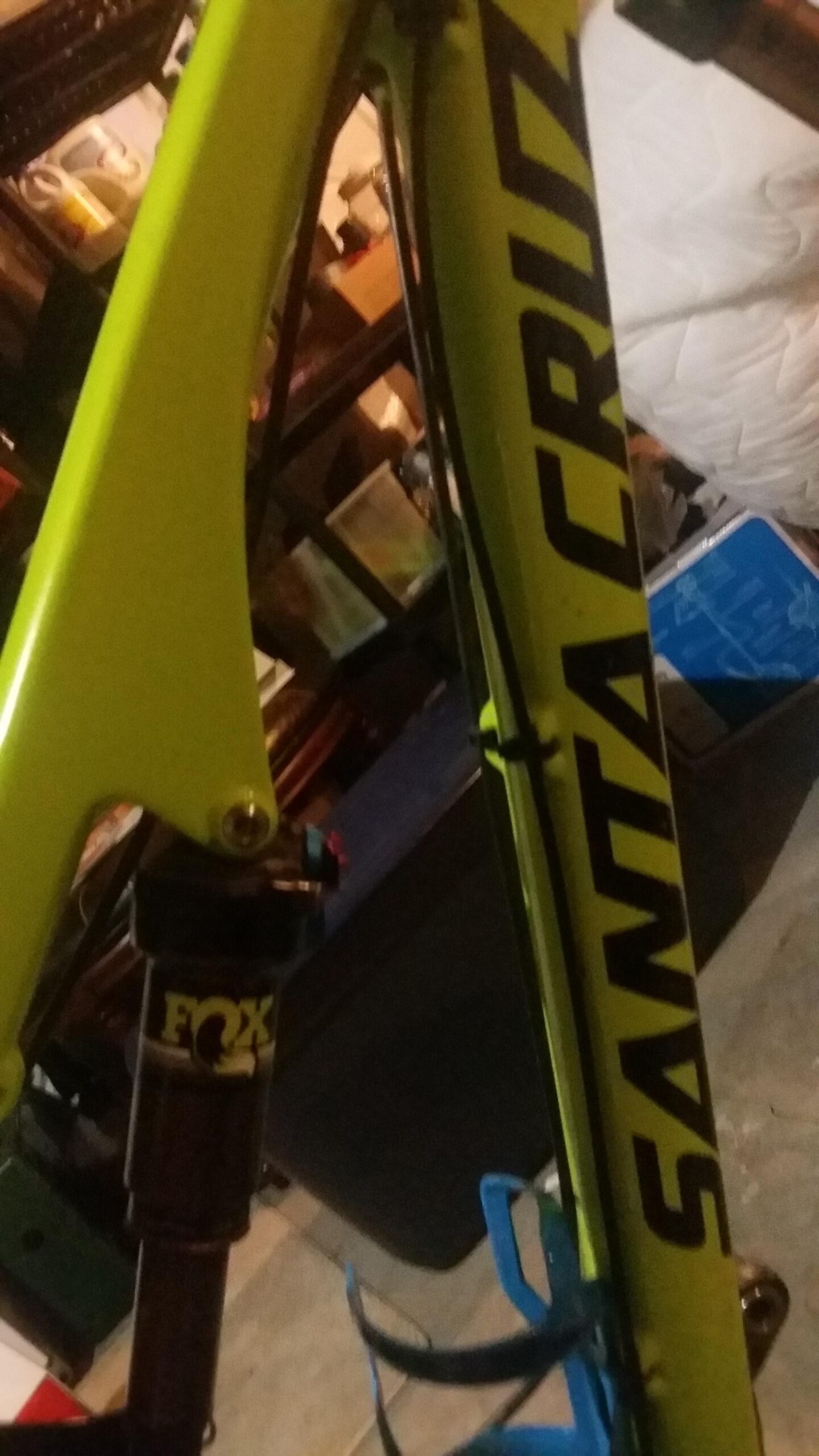 Santa Cruz Bronson: Close-up of a bright green mountain bike frame with the "Santa Cruz" logo prominently displayed. The image also features a Fox suspension component. The background shows a cluttered garage environment.