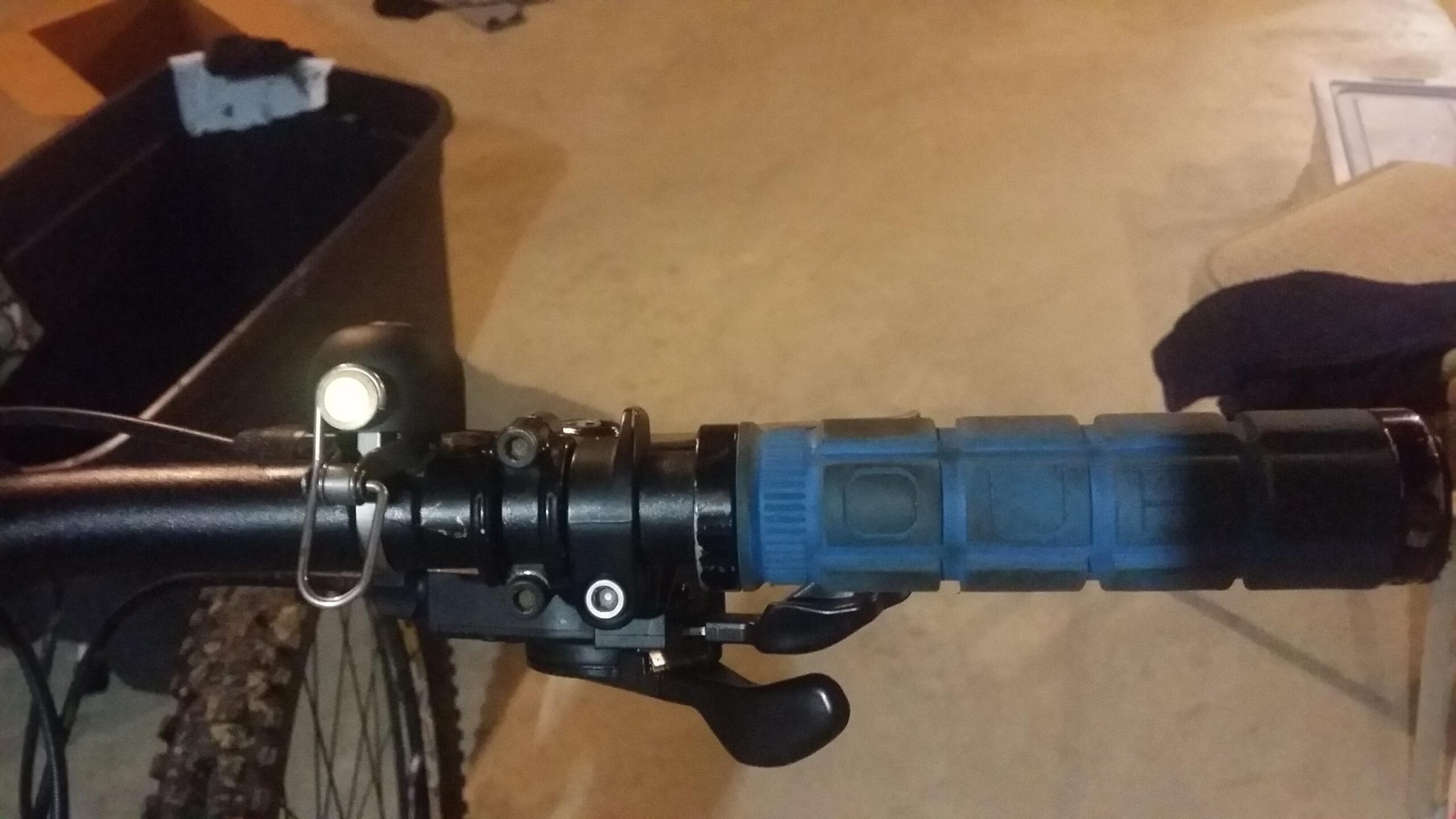 Santa Cruz Bronson: Close-up view of a bicycle handlebar featuring a black grip, a bicycle bell, and a gear shift lever. The background is a garage setting with a black storage bin and scattered clothing items.