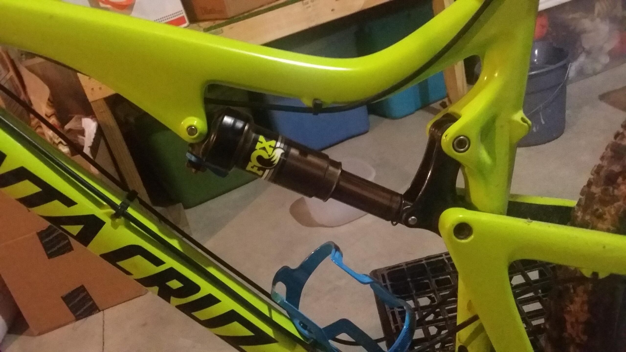 Santa Cruz Bronson: A close-up view of a bright green mountain bike frame, showcasing the rear suspension and shock absorber. The bike features a black Fox shock, and a blue water bottle cage is attached to the frame. The background includes indistinct objects, suggesting a garage or workshop setting.