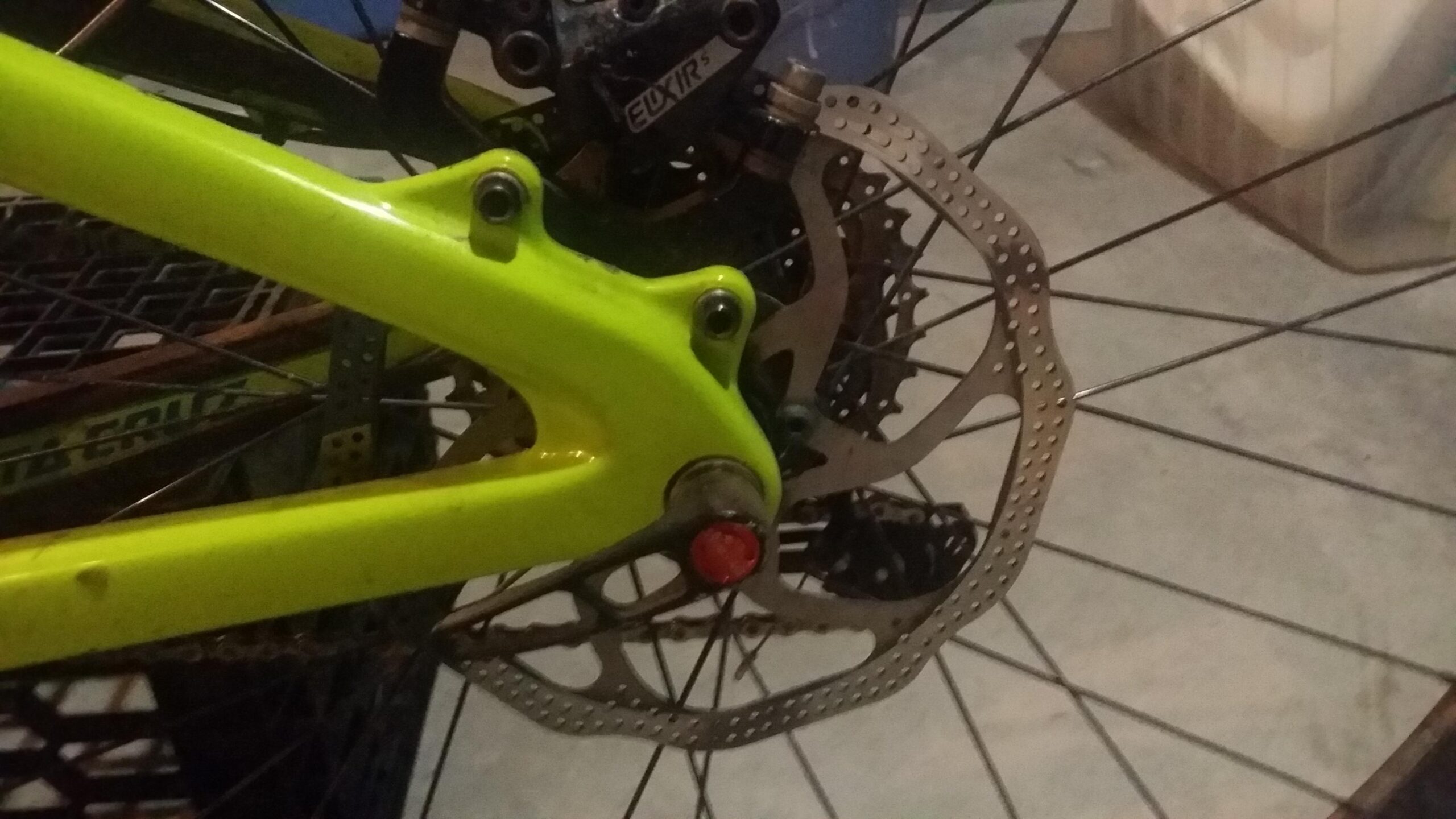 Santa Cruz Bronson: Close-up view of a bicycle's rear wheel assembly, featuring a bright green frame and a disc brake system. The image highlights the brake rotor and the caliper, along with a red bolt or cap near the hub. The background shows a textured surface, suggesting a garage or workshop setting.