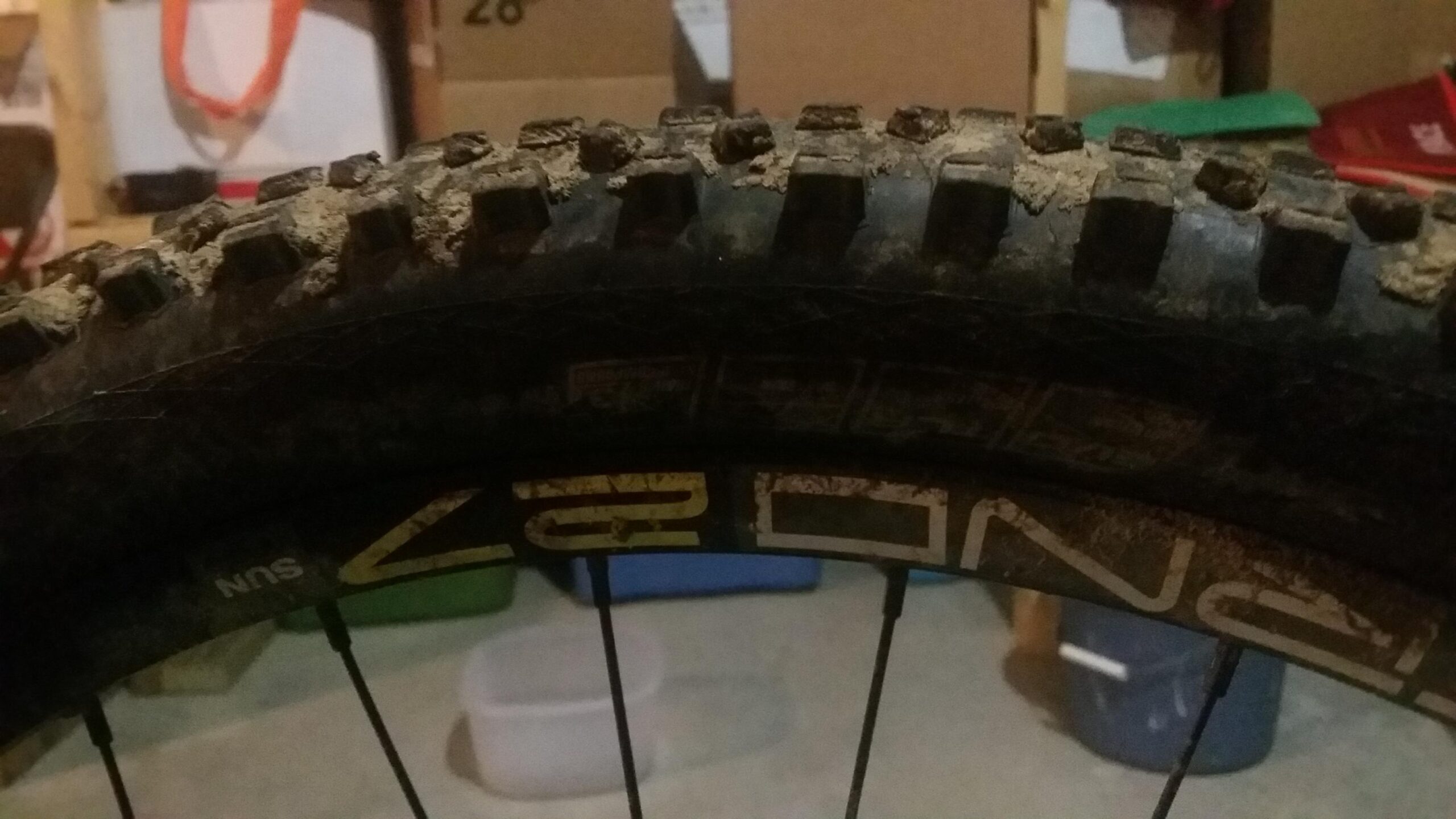 Schwalbe Hans Dampf: Close-up view of a muddy mountain bike tire, showcasing the tread pattern and a portion of the bike rim. The background features blurred boxes and storage items.