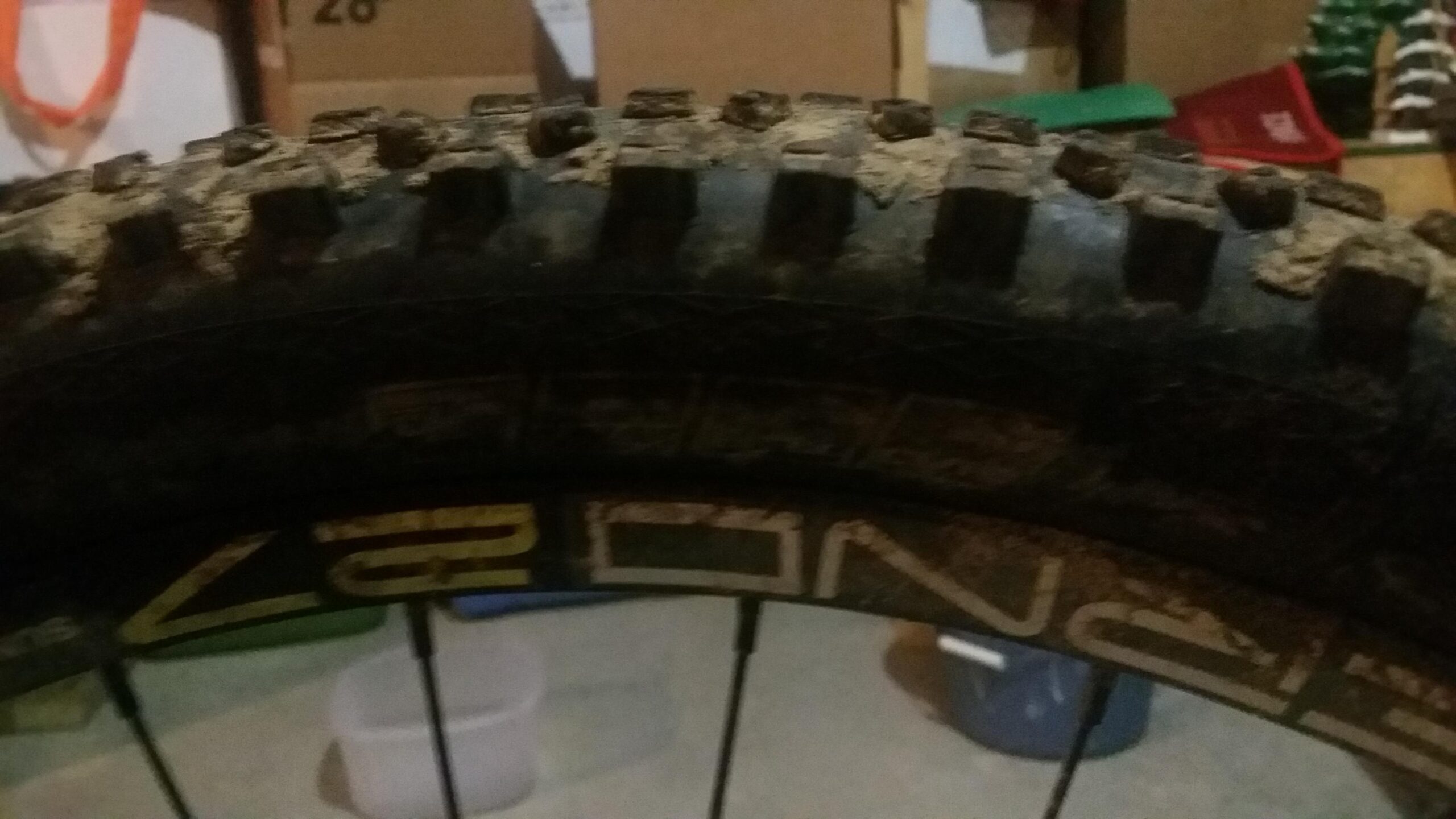 Schwalbe Hans Dampf: Close-up image of a muddy mountain bike tire, showcasing the tread pattern and sidewall markings. In the background, there are blurred boxes and storage containers, indicating a garage or storage area.