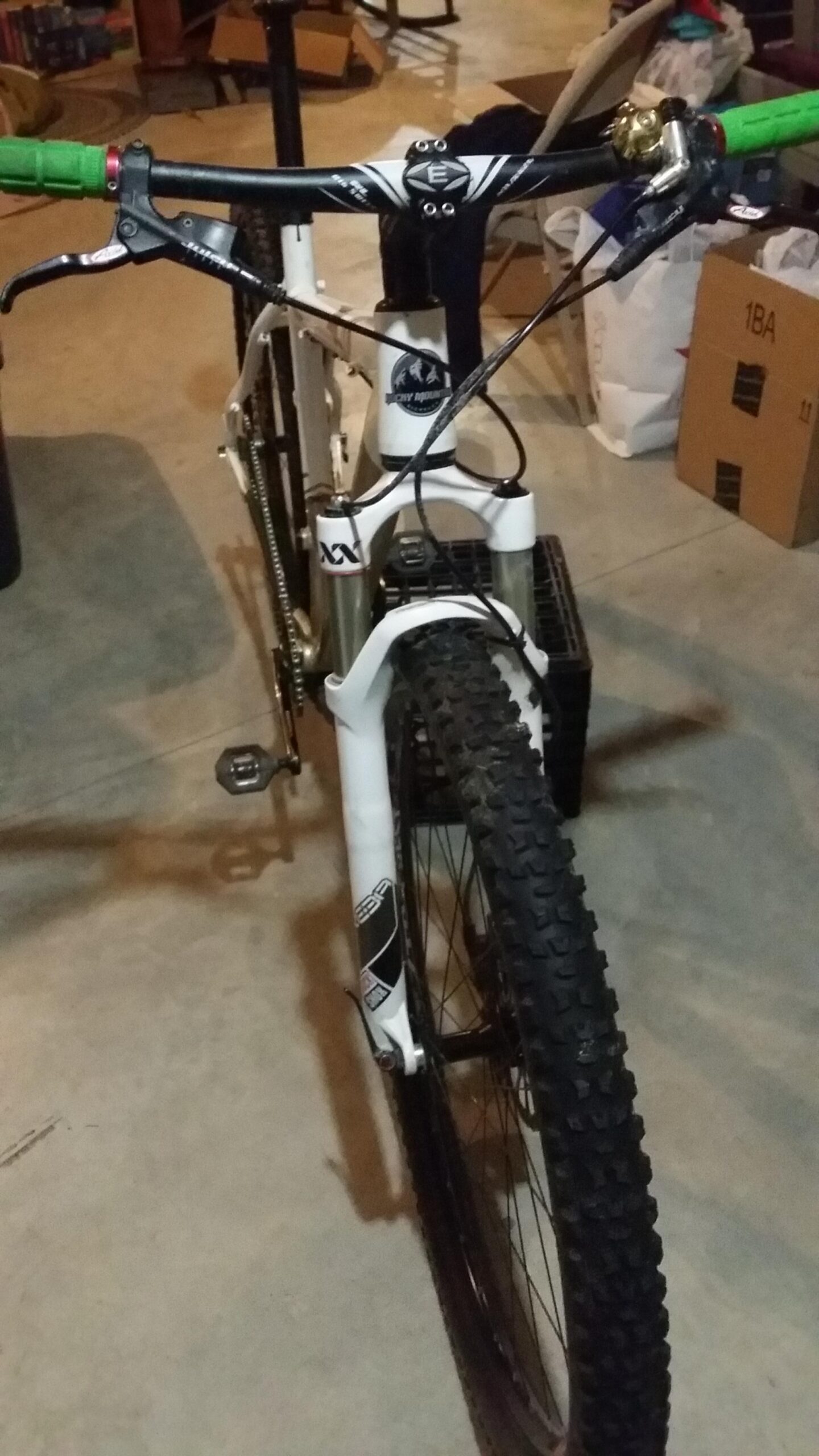 Rocky Mountain Vertex 990 rsl 2013: A white mountain bike positioned in a garage, showing the front view with green handle grips, a bell, and a sturdy front tire with deep tread. The background features various items, including boxes and a chair.