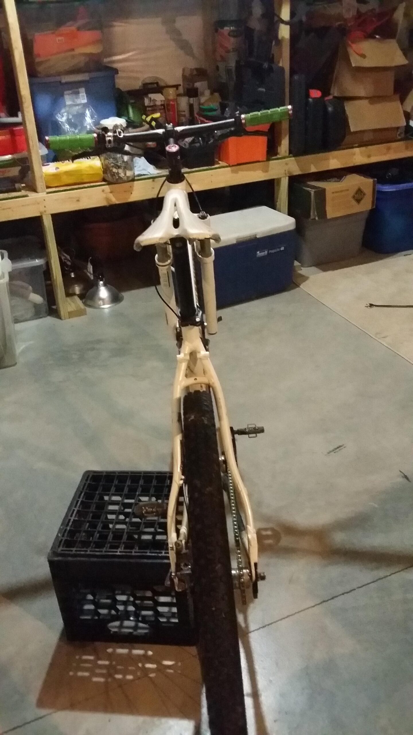 Rocky Mountain Vertex 990 rsl 2013: A rear view of a bicycle positioned on a black plastic crate, set against a backdrop of a cluttered garage. The bike has a white frame and green handlebar grips, with items and storage boxes visible in the background. The garage floor is concrete, and there are various tools and containers stored on shelves.