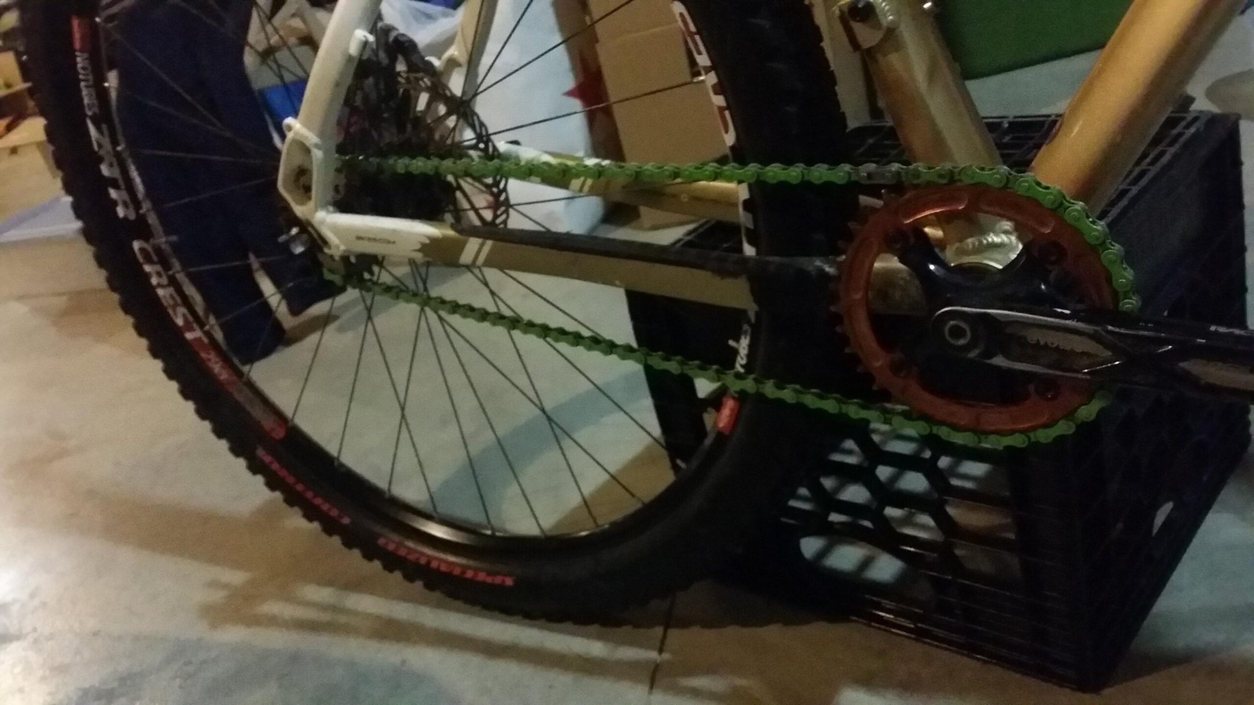 Rocky Mountain Vertex 990 rsl 2013: Image of a bicycle's drivetrain, featuring a green chain running over a gold and black chainring, with a close-up of the pedal mechanism. The background includes a black crate and other biking equipment, illuminated in low light conditions.