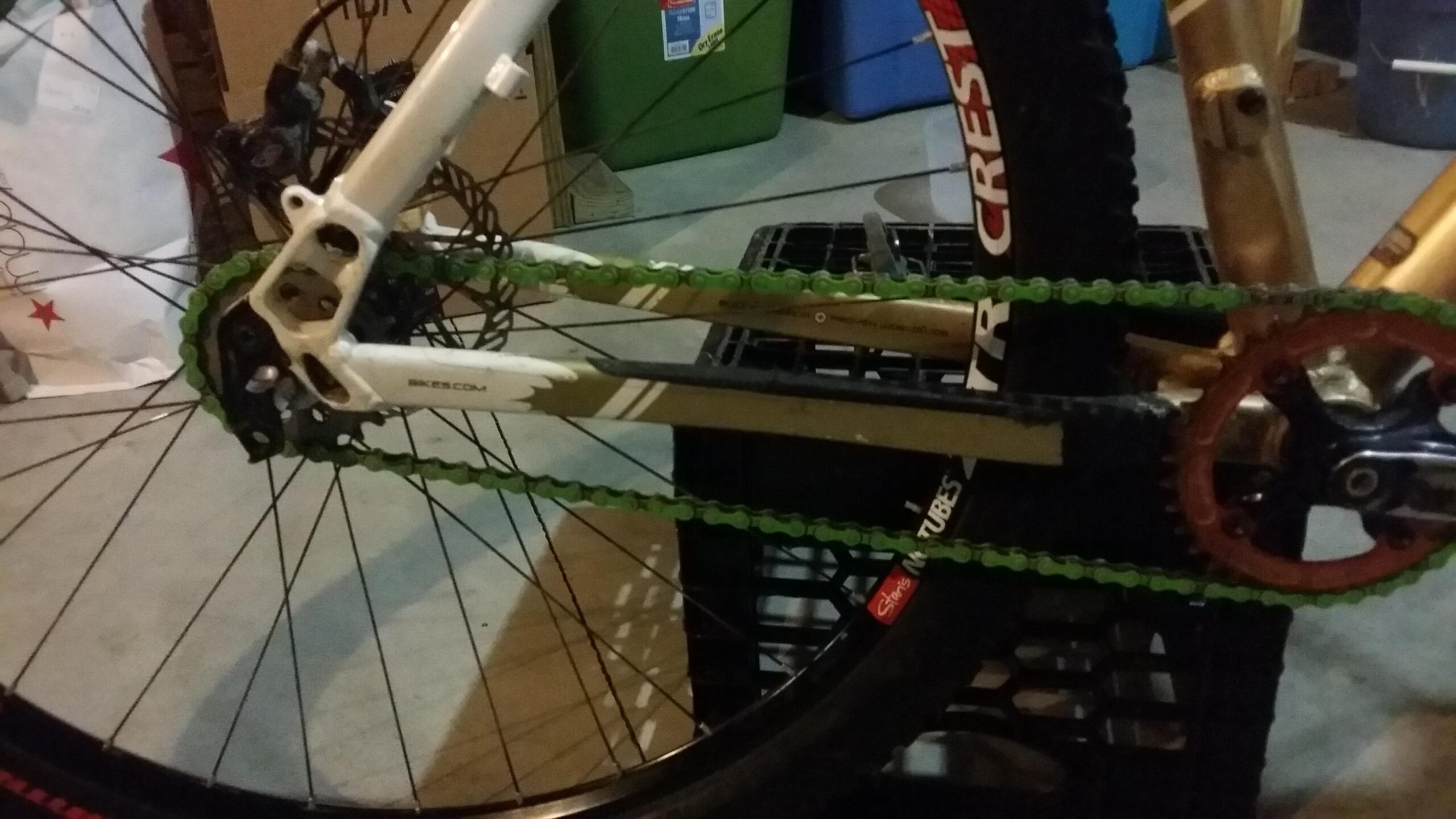 Rocky Mountain Vertex 990 rsl 2013: Close-up view of a bicycle's drivetrain, featuring a green chain, a red sprocket, and a white frame. The bike is positioned on a black crate, with various items partially visible in the background.