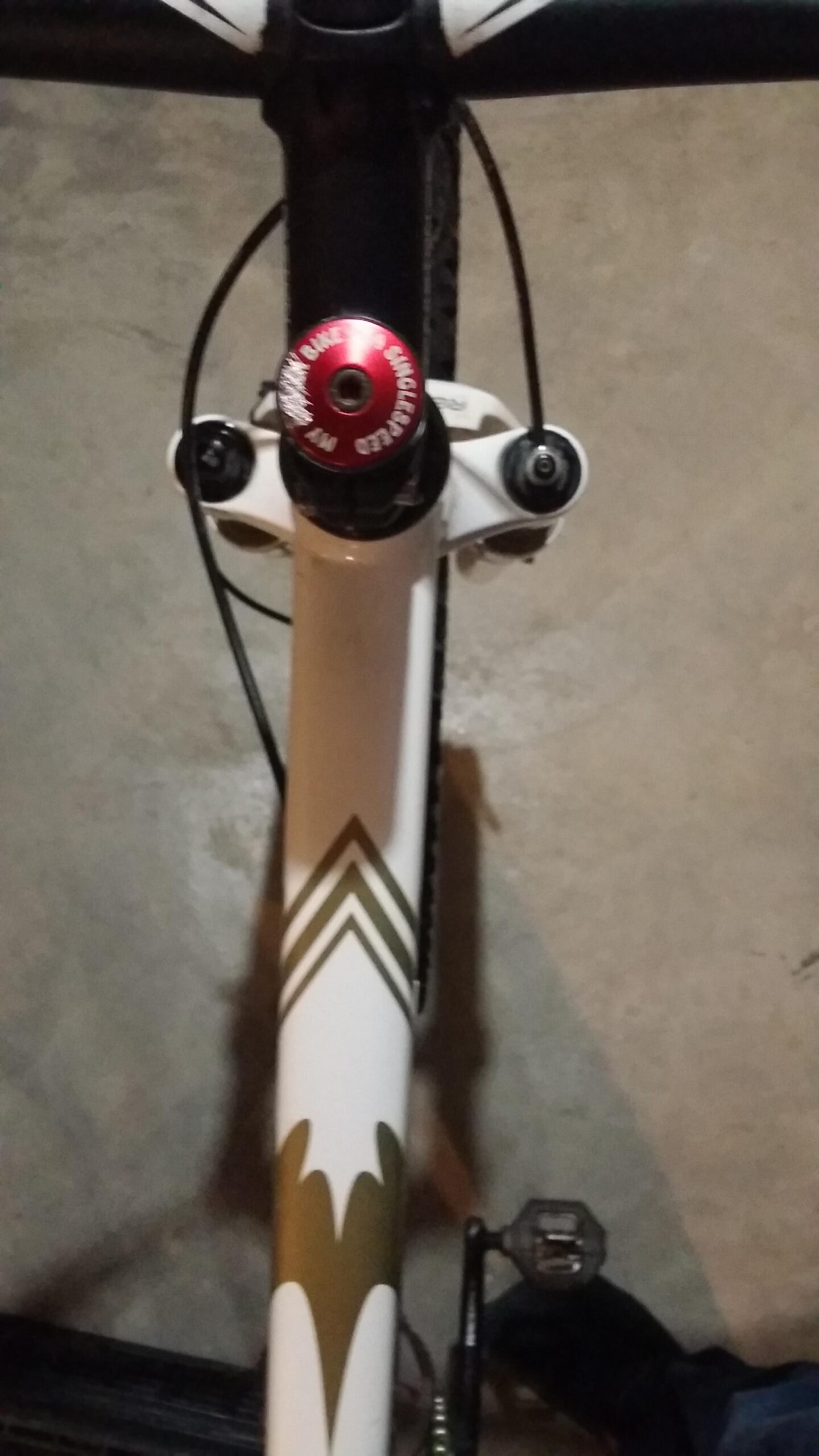 Rocky Mountain Vertex 990 rsl 2013: A close-up view of a bicycle's handlebars and stem, featuring a red headset cap and a white frame with a green and gold geometric design. The bike is positioned on a concrete floor, with part of a pedal visible on the right side.