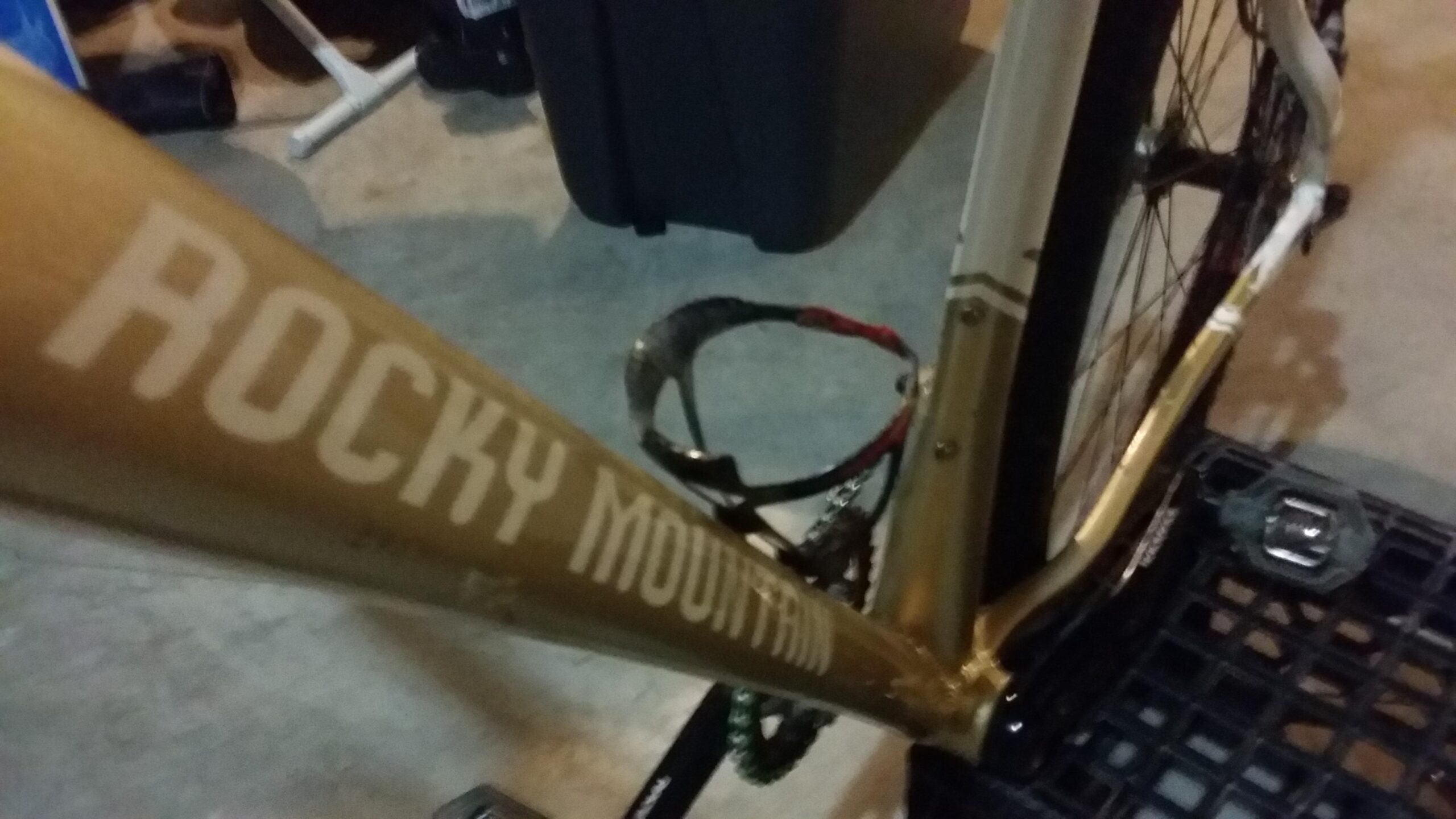 Rocky Mountain Vertex 990 rsl 2013: Close-up image of a bicycle frame featuring the text "ROCKY MOUNTAIN" in bold lettering. The frame is gold in color and is positioned on a textured surface. A pedal and part of the bike's drivetrain are also visible in the background.