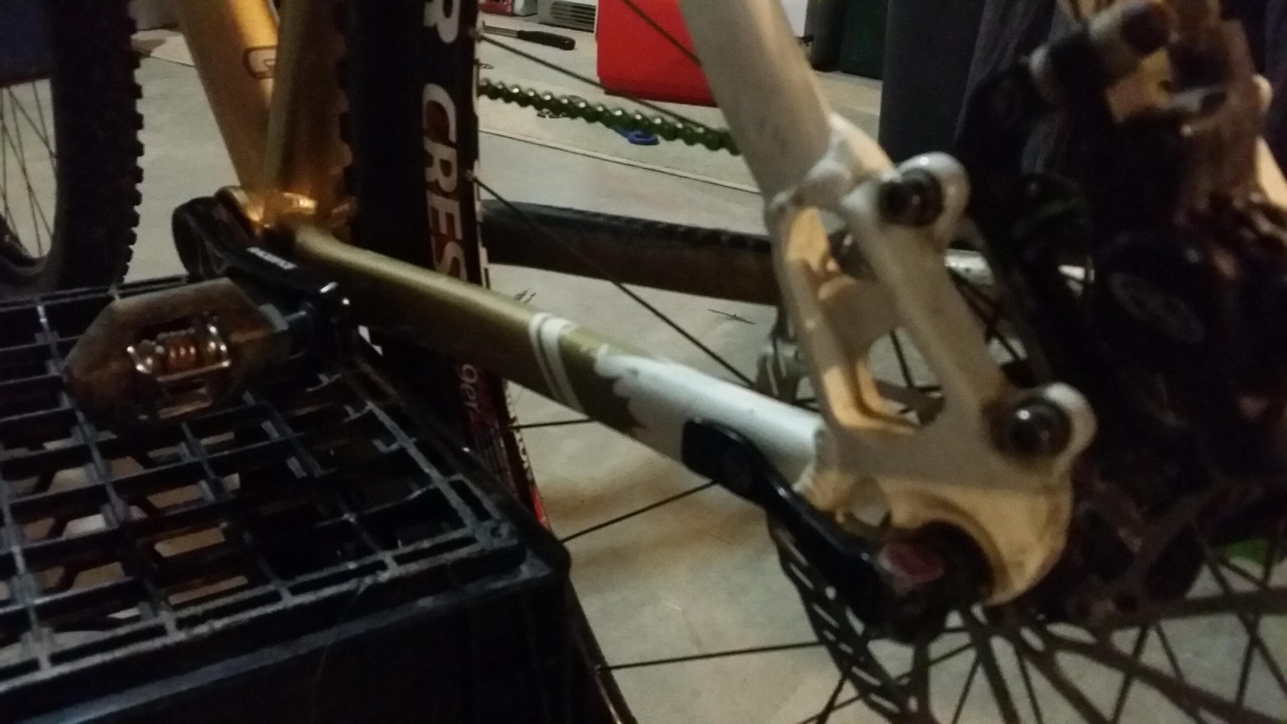 Rocky Mountain Vertex 990 rsl 2013: Close-up image of a bicycle's crank and pedal assembly, featuring a golden frame and a black pedal attached to a crate. The background includes elements of a garage setting, with tools and bike components partially visible. The focus highlights the mechanical details and construction of the bike's drivetrain.