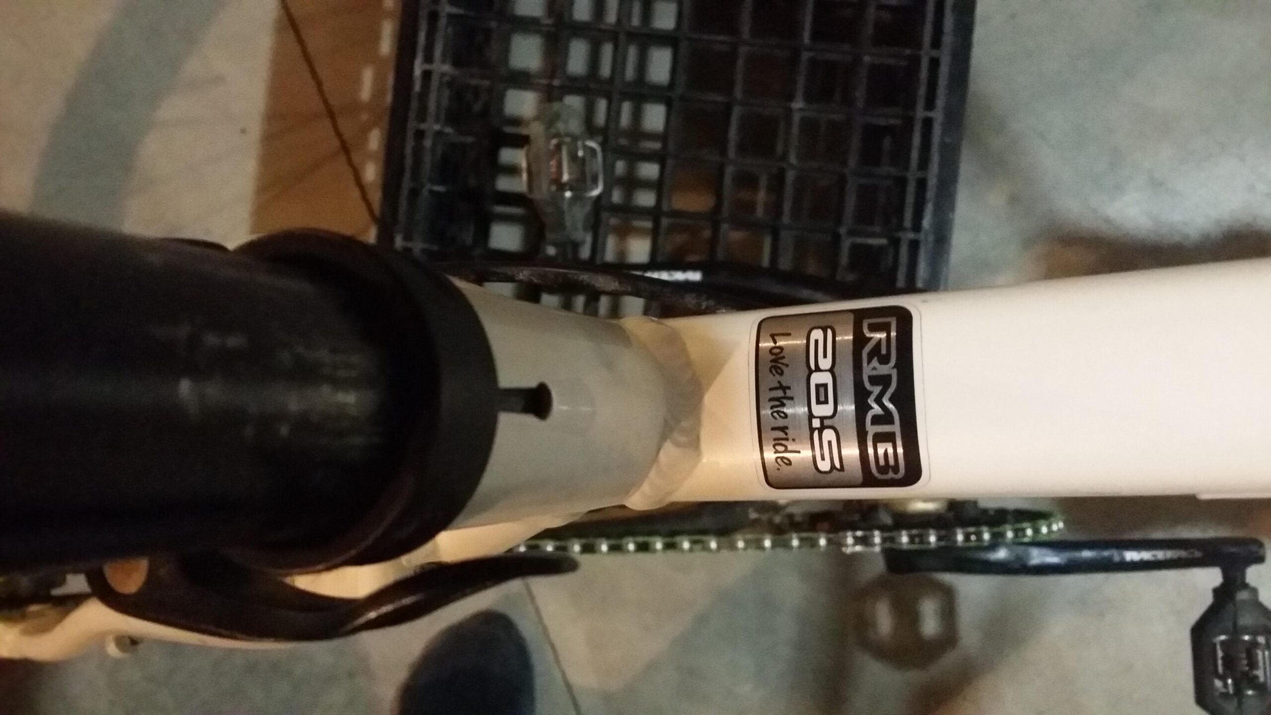 Rocky Mountain Vertex 990 rsl 2013: Close-up image of a bike frame featuring a "RMB 20.S" decal with the phrase "Love the ride." The frame is black and white, showing part of the bike's crank and chain. A plastic crate is visible in the background, along with a section of the ground.