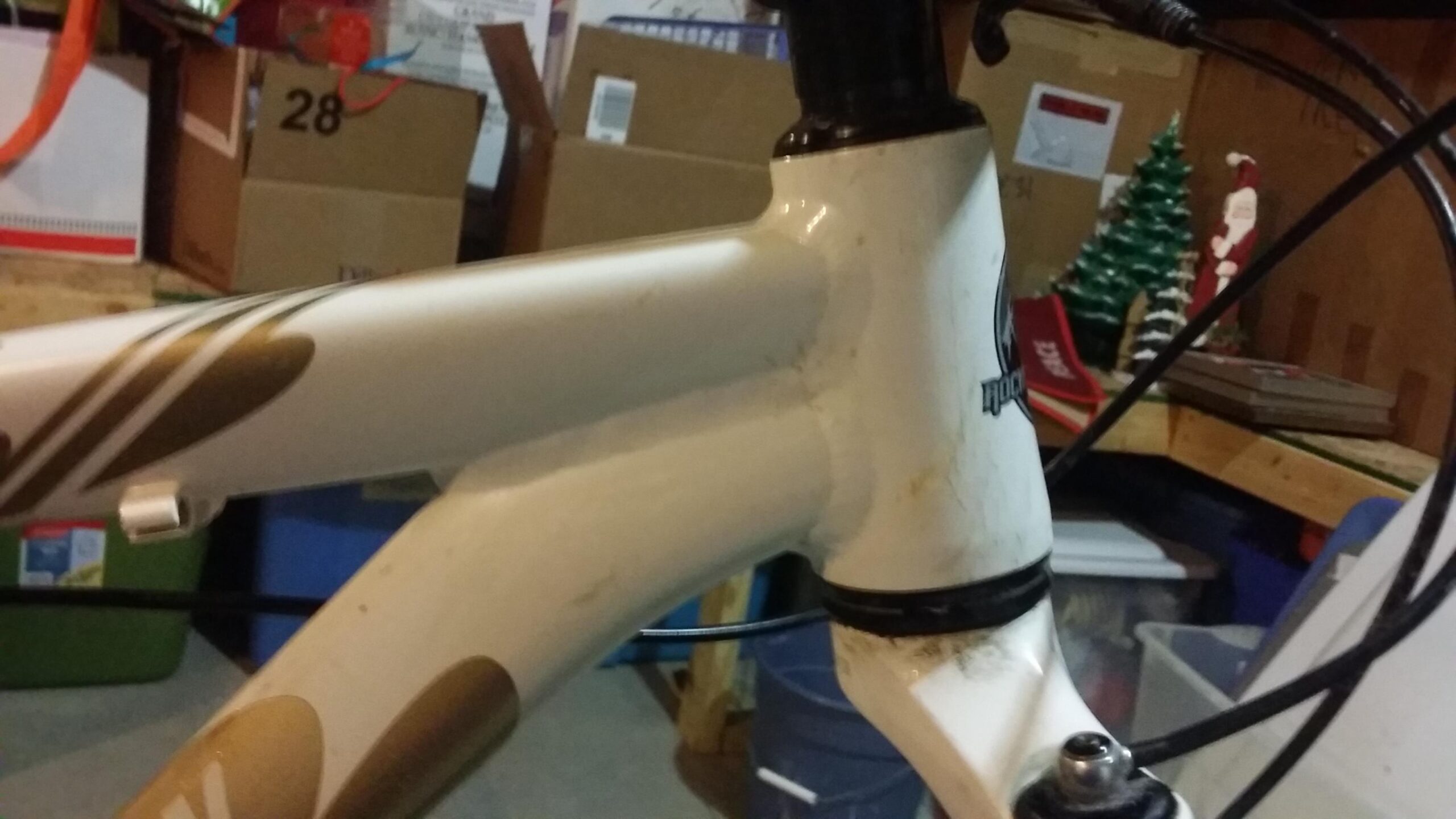 Rocky Mountain Vertex 990 rsl 2013: Close-up image of a bicycle's frame at the junction of the top tube and head tube. The frame is primarily white with gold accents and shows signs of dirt and wear. In the background, storage boxes and holiday decorations, including a small Christmas tree and Santa figure, are visible.