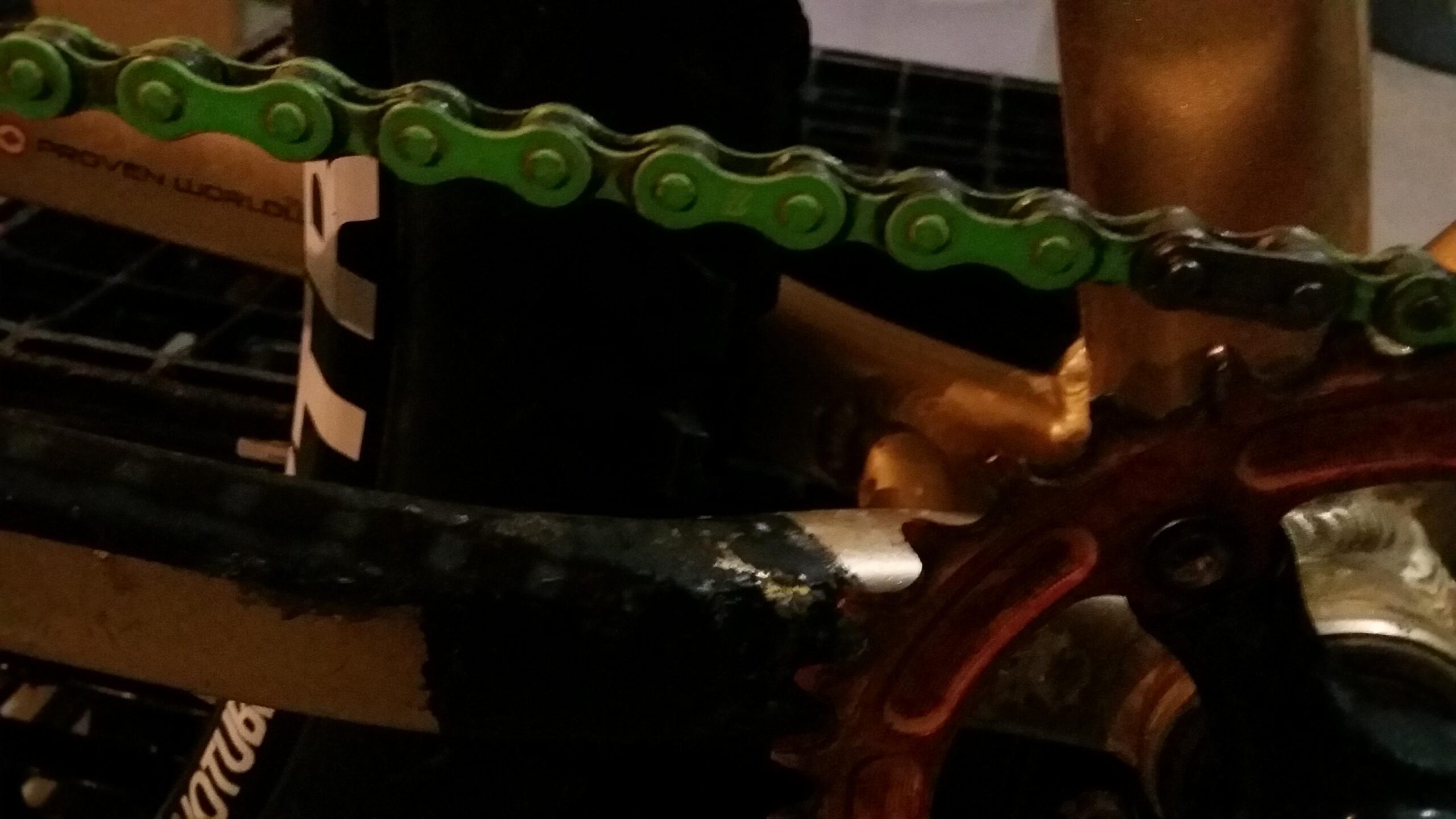 Rocky Mountain Vertex 990 rsl 2013: A close-up view of a bicycle chain, featuring a vibrant green chain composed of interconnected links. The chain is positioned against the bicycle's crankset, which appears to be partially worn and shows signs of use. The background is dimly lit, highlighting the texture and details of the bike’s components.