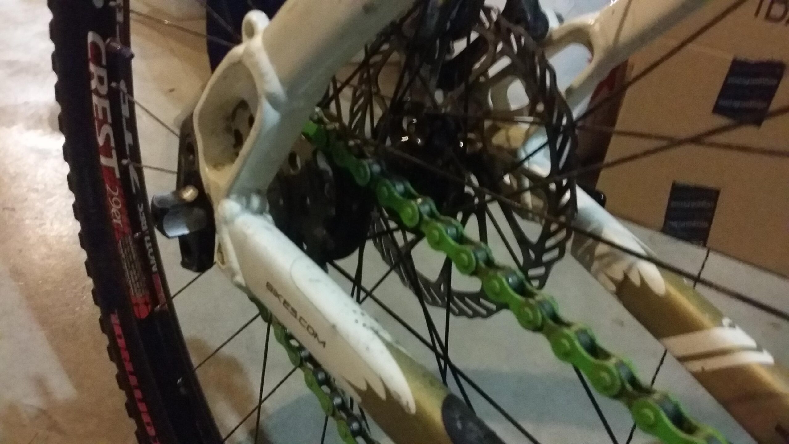 Rocky Mountain Vertex 990 rsl 2013: Close-up view of a bicycle's rear drivetrain, showcasing a green chain, sprocket, and disc brake. The bike frame is primarily white with some dirt visible, and part of the tire with black tread is in the foreground. Background includes a blurred carton box.