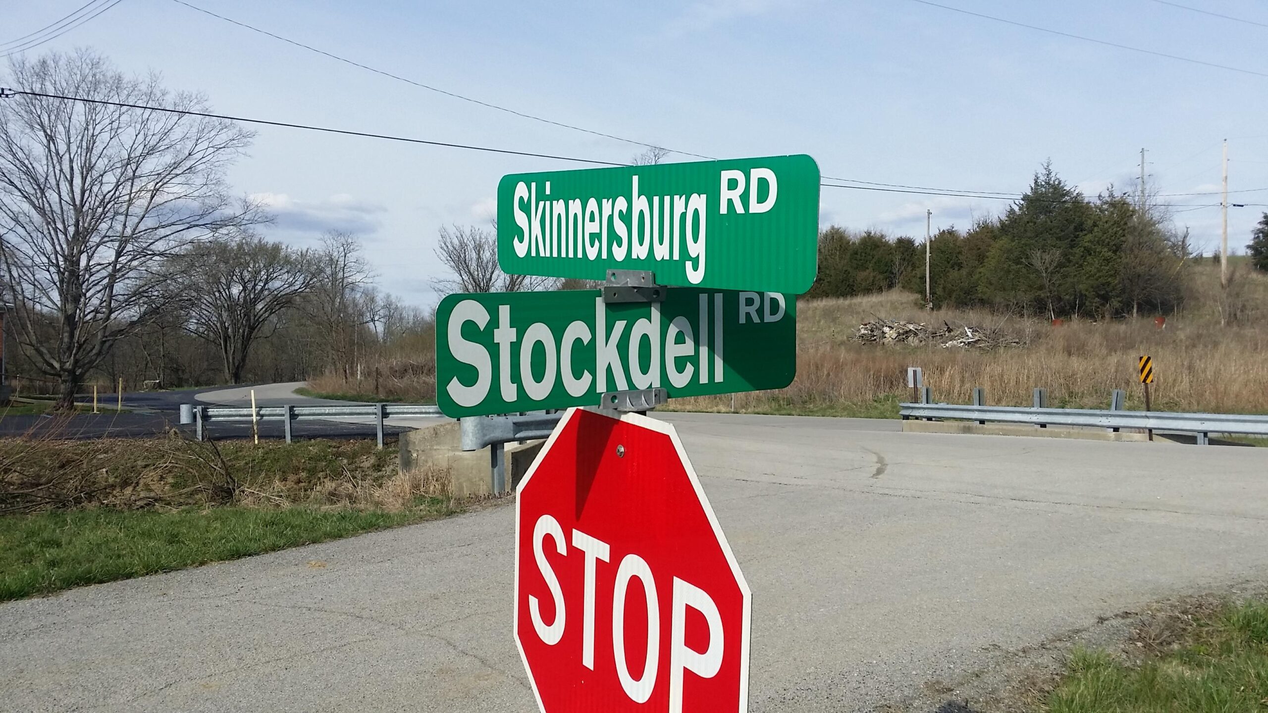 A street intersection sign showing "Skinnerburg Rd" and "Stockdell Rd," with a visible red stop sign in the foreground. The scene features a rural landscape with sparse trees and a clear sky. Skullbuster mountain bike trail.