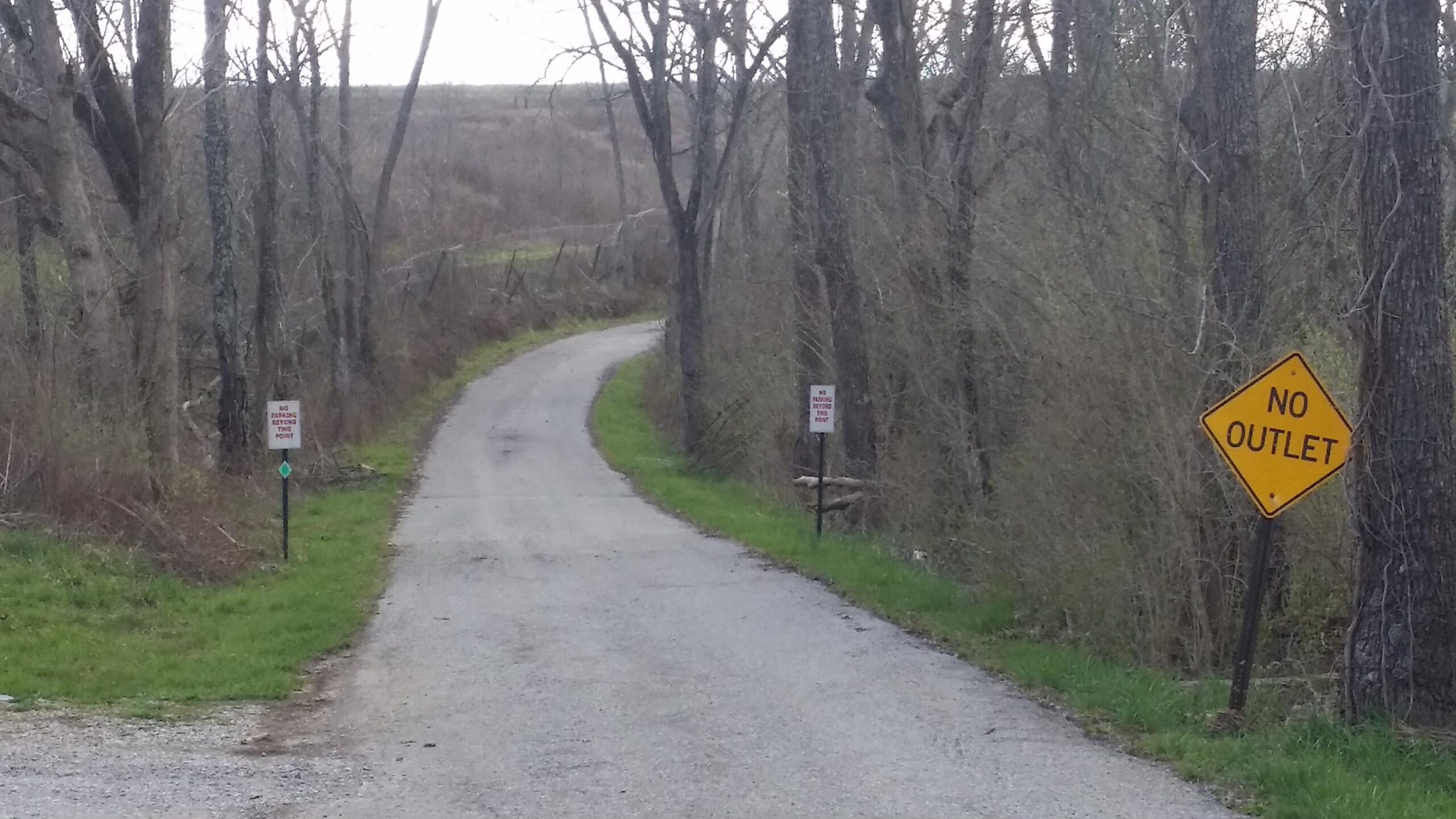 A winding gravel road surrounded by bare trees and dense brush. On the right side of the road, a yellow "No Outlet" sign is visible. Two additional signs are posted on the left, indicating "No Parking." The scene appears peaceful and secluded, with a cloudy sky in the background. Skullbuster mountain bike trail.