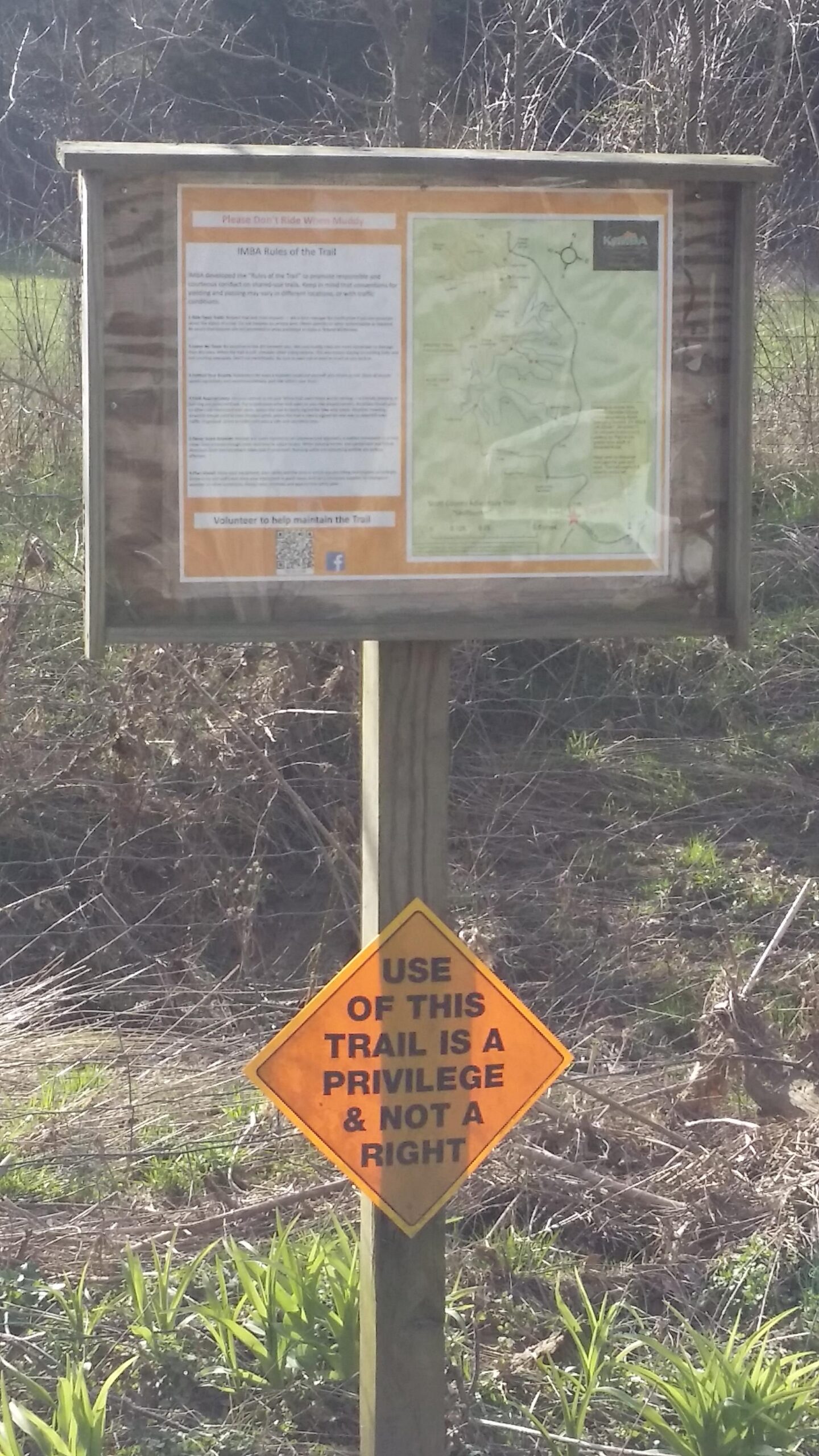 A trail information sign with a map and rules for trail use, accompanied by a warning sign that states, "USE OF THIS TRAIL IS A PRIVILEGE & NOT A RIGHT." The background features uneven terrain with sparse vegetation. Skullbuster mountain bike trail.
