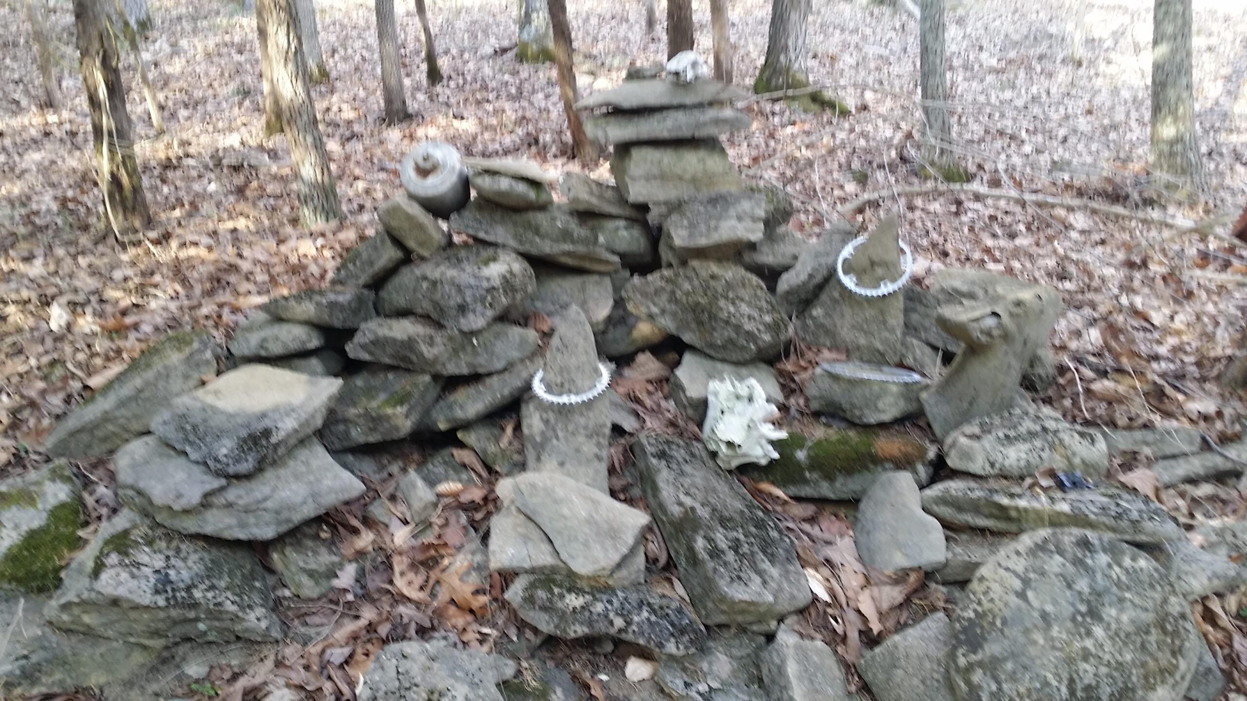 A stone structure composed of various rocks, some adorned with what appears to be circular decorations, situated among fallen leaves and trees in a forested area. A skull and a pot-like object are also visible among the stones, creating an organic and possibly decorative arrangement. Skullbuster mountain bike trail.