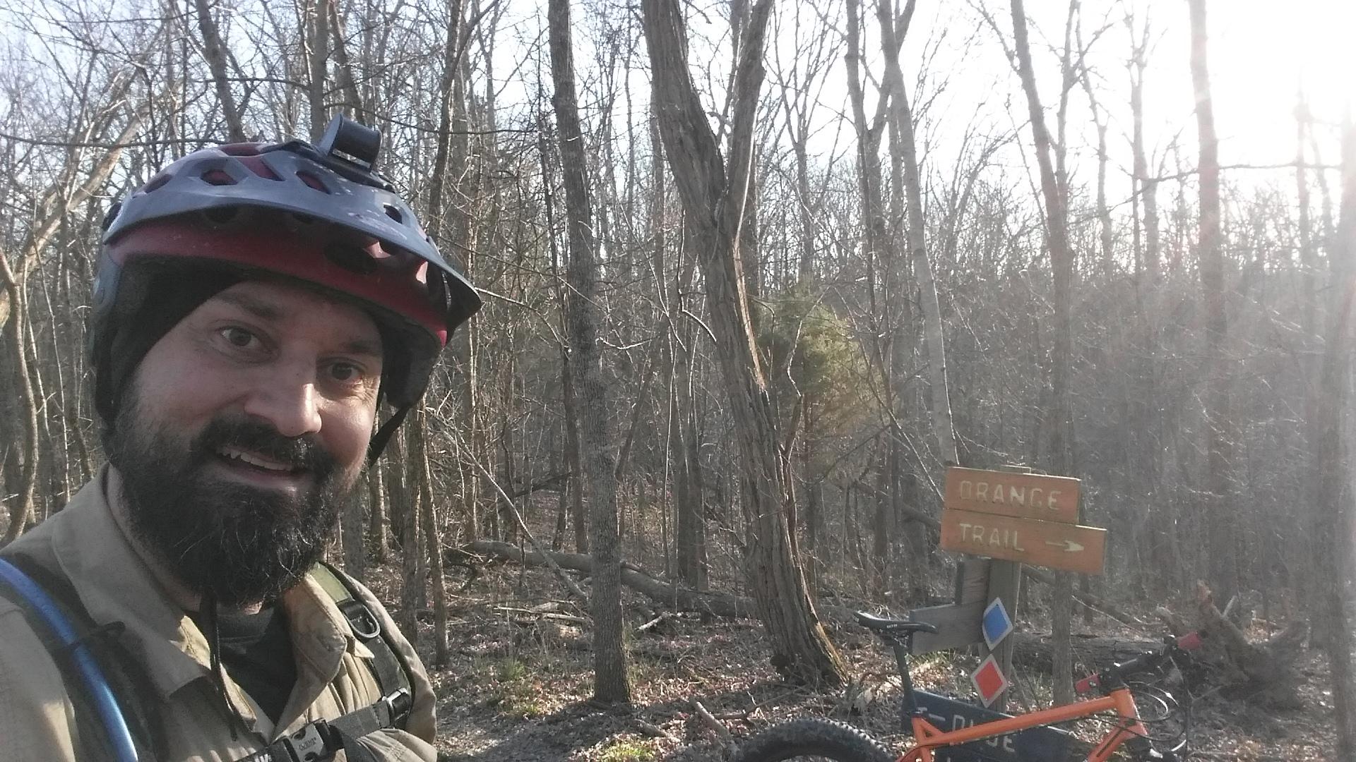 A person wearing a bicycle helmet and a backpack smiles at the camera while standing on a dirt trail surrounded by trees. In the background, there is a wooden sign indicating the direction of the "Orange Trail." A bicycle can be seen leaning against a post near the sign. The scene is illuminated by natural sunlight filtering through the branches. Skullbuster mountain bike trail.