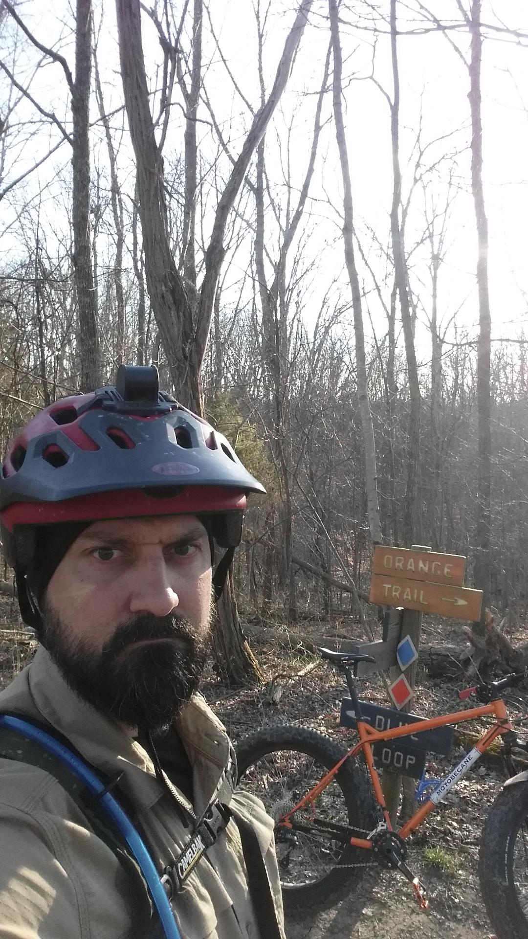 A person wearing a helmet and winter attire stands in a wooded area, taking a selfie. In the background, there is a sign indicating the "Orange Trail" direction and an orange mountain bike parked beside it. The scene is set during the day with bare trees, suggesting early spring or late winter. Skullbuster mountain bike trail.