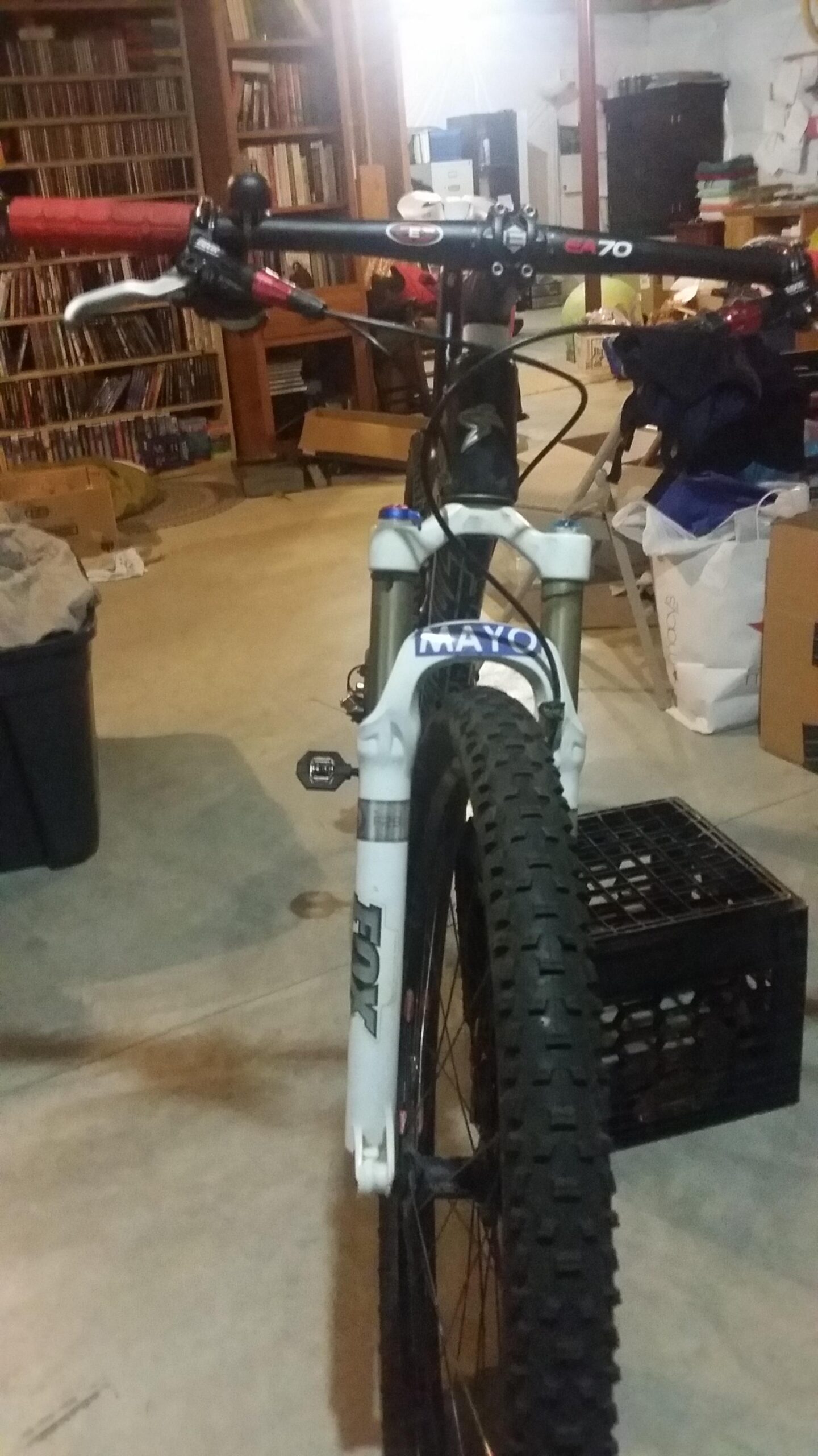 Santa Cruz Tallboy Carbon: A mountain bike viewed from the front, showcasing its handlebars with red grips and brake levers, a white front fork, and a knobby tire. The background features shelves with books and various items, suggesting a cluttered indoor space, likely a garage or basement.