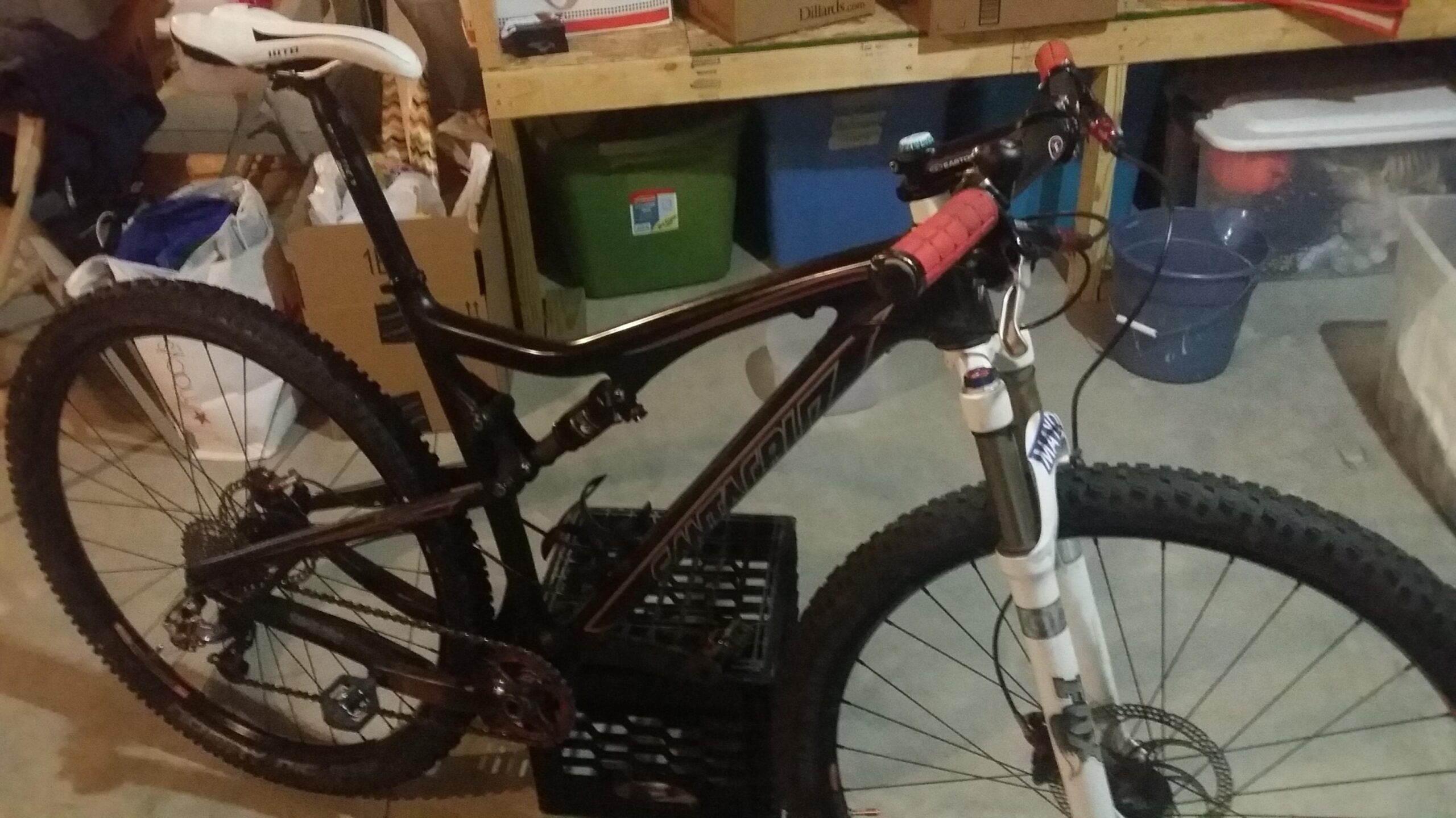 Santa Cruz Tallboy Carbon: A mountain bike positioned on a crate in a garage, featuring a black frame with red handlebar grips and a white saddle. Various storage items and boxes are visible in the background.